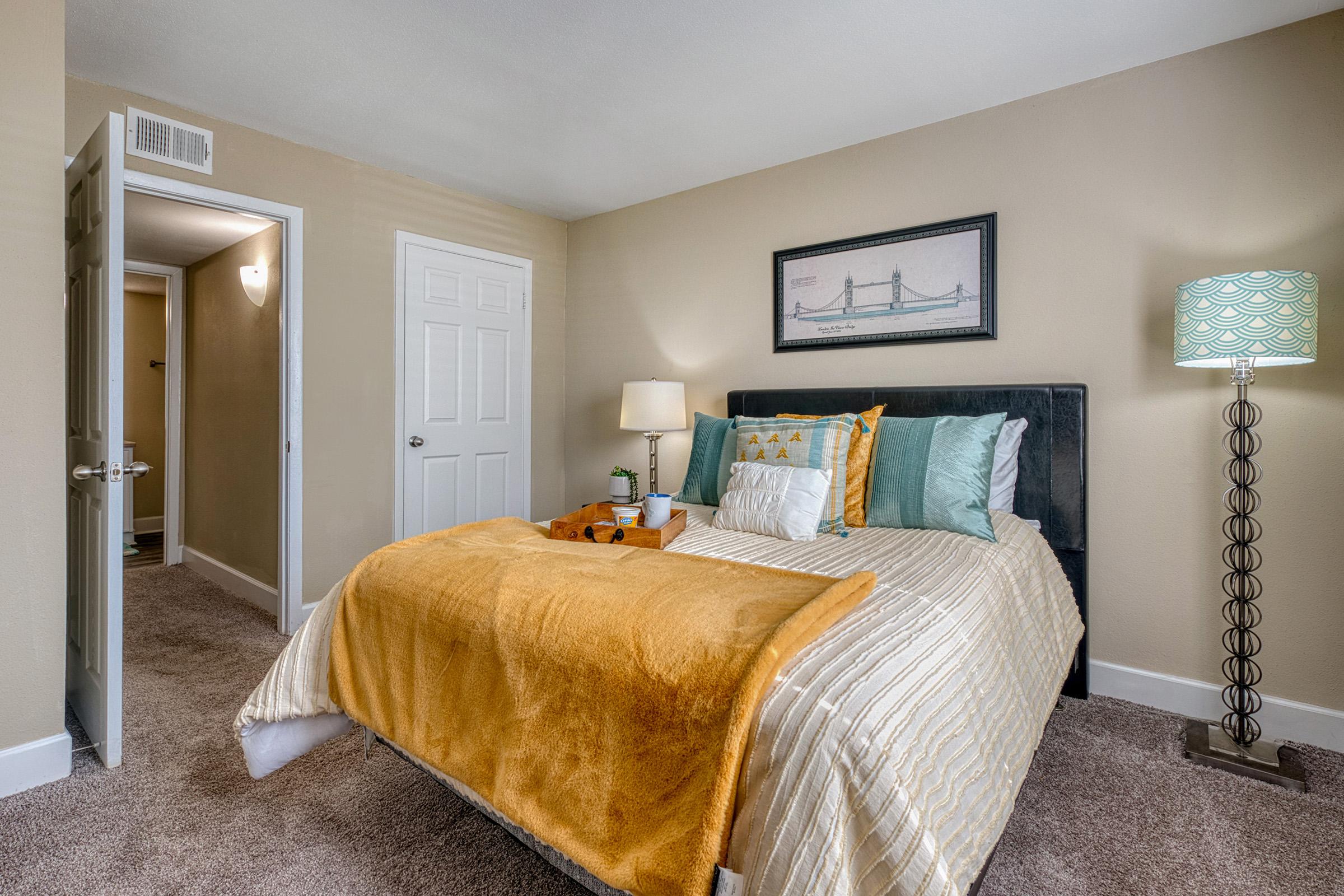 Cozy bedroom featuring a neatly made bed with orange and teal bedding, a bedside lamp, and decorative pillows. A framed picture of a bridge is hanging on the wall. Light brown carpet and a door leading to a hallway are visible, creating a warm and inviting atmosphere.