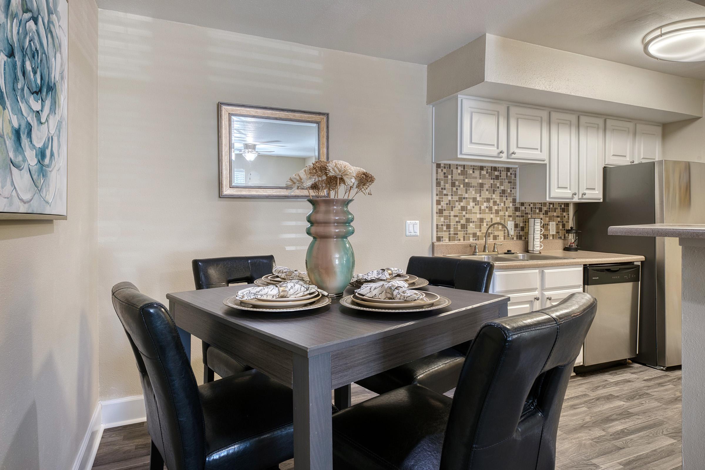 A dining area featuring a wooden table set for four with decorative plates and napkins. A stylish vase with dried flowers sits at the center. The background includes a modern kitchen with white cabinets, a mosaic backsplash, and stainless steel appliances. A mirror reflects part of the space, enhancing the light and openness.