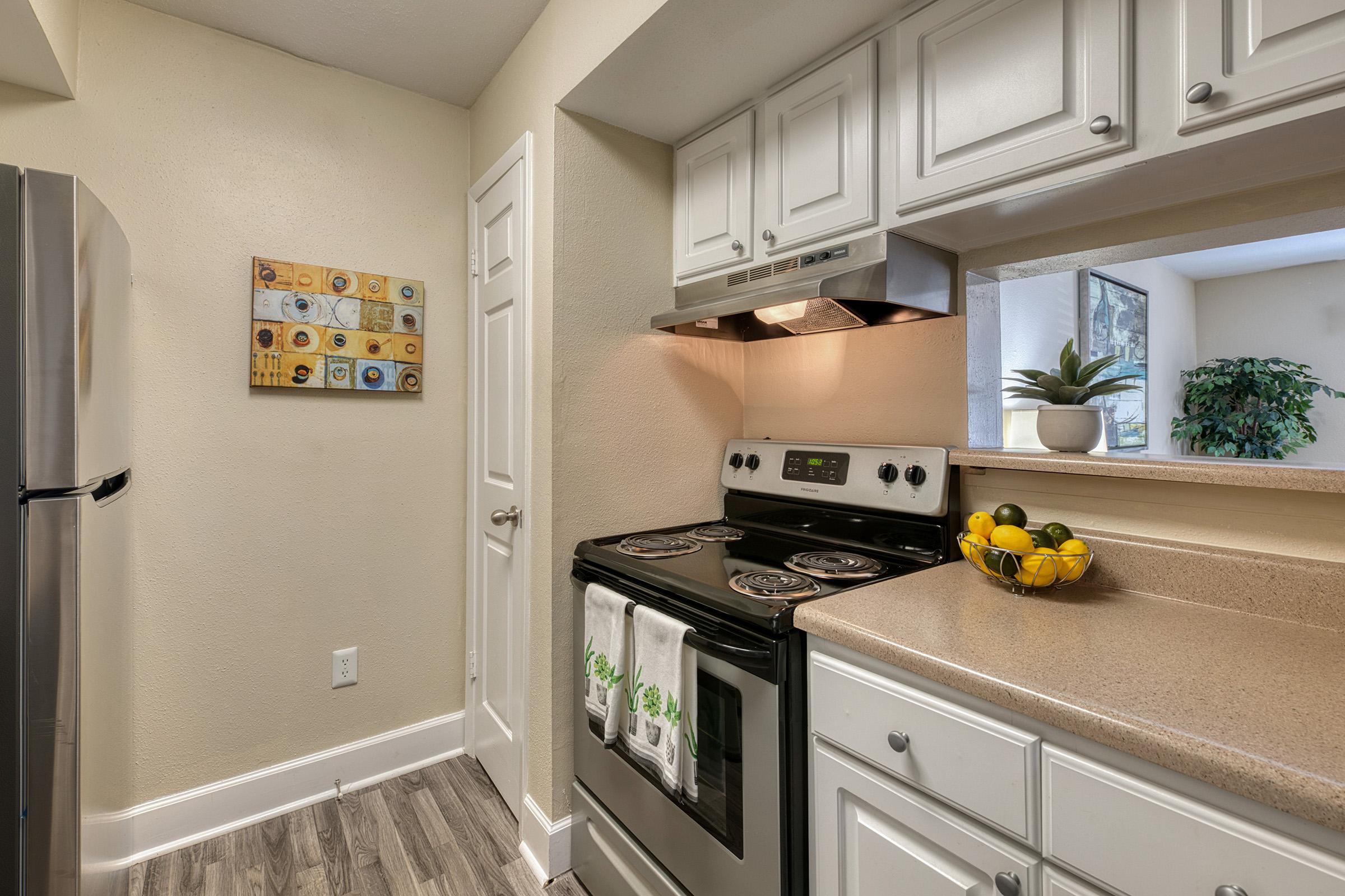 Modern kitchen featuring white cabinetry, stainless steel refrigerator, and a black stove with a range hood. A bowl of colorful fruits sits on the countertop, complemented by a decorative wall art piece. Natural light illuminates the space, highlighting the neutral color scheme and clean design.