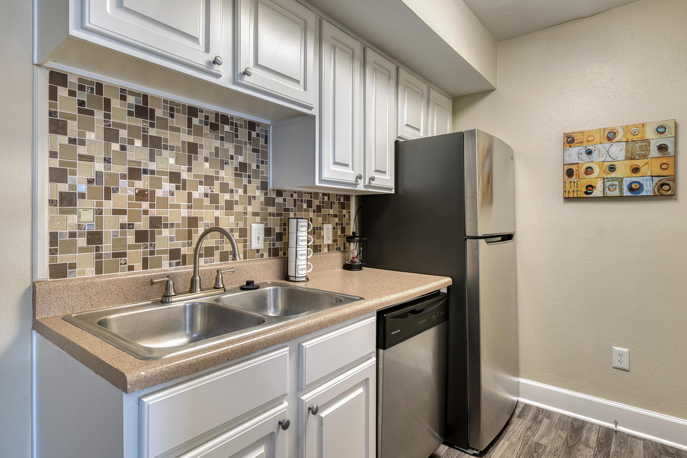 A modern kitchen featuring white cabinetry, a double sink with a chrome faucet, and stainless steel appliances, including a refrigerator and dishwasher. The backsplash consists of decorative tiles in various earth tones, and there are kitchen essentials like a coffee maker and a towel on the counter.