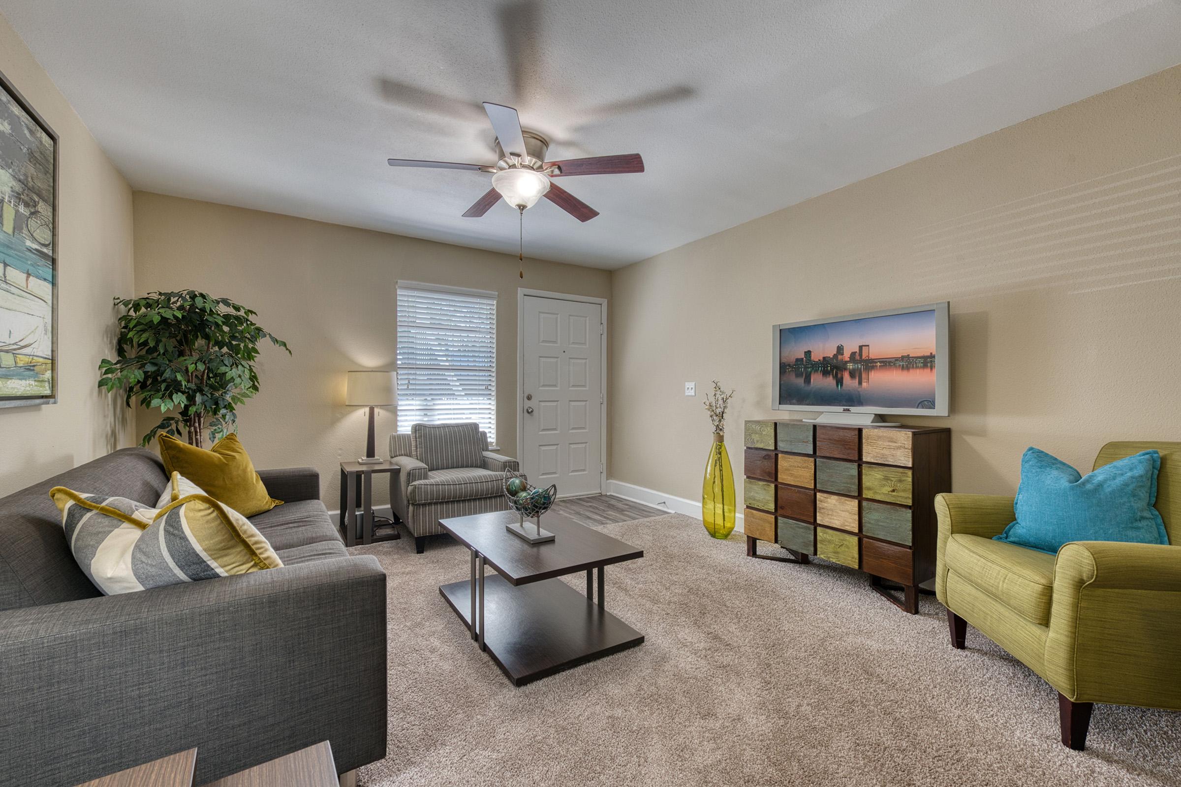 A cozy living room featuring a gray sofa with colorful accent pillows, a wooden coffee table, a decorative TV stand, and a green armchair. The space includes a fan on the ceiling, potted plant, and large windows that bring in natural light, creating a warm and inviting atmosphere.
