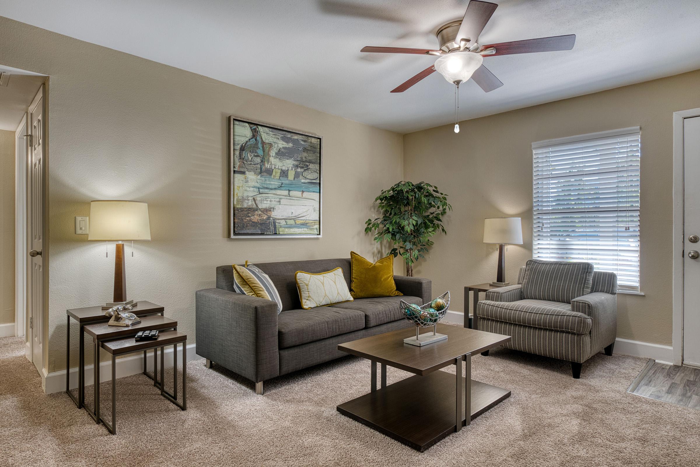 A cozy living room featuring a gray sofa with yellow accent pillows, a stylish armchair, a wooden coffee table, and contemporary side tables. Soft lighting from two lamps and a ceiling fan create a warm atmosphere. A wall art piece and a potted plant add decorative touches to the space.