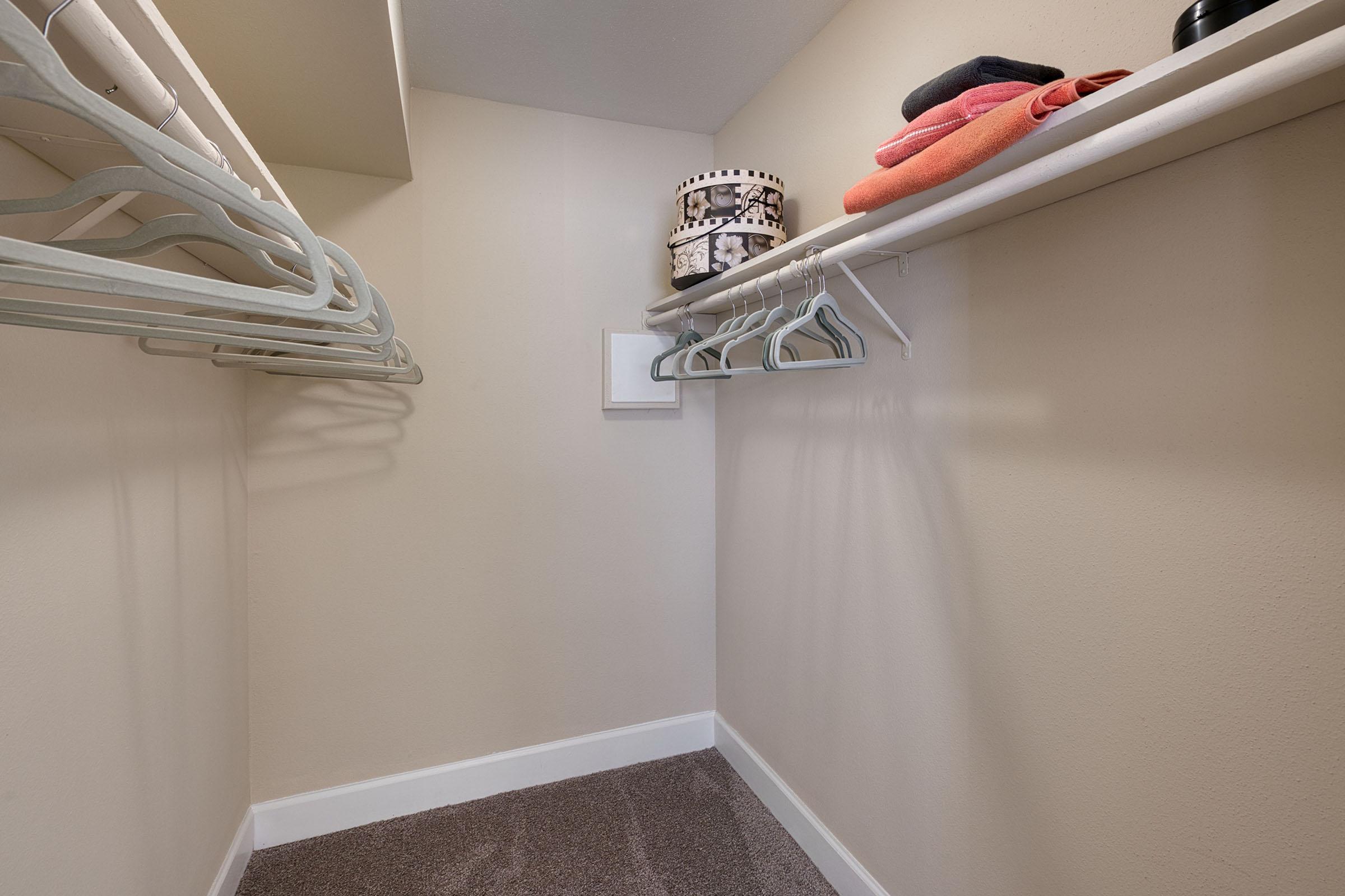 A well-organized closet featuring several white hangers on a rod, neatly arranged colorful towels stacked on a shelf, and a decorative storage basket. The walls are painted a soft beige, and the floor is carpeted in a light brown shade.