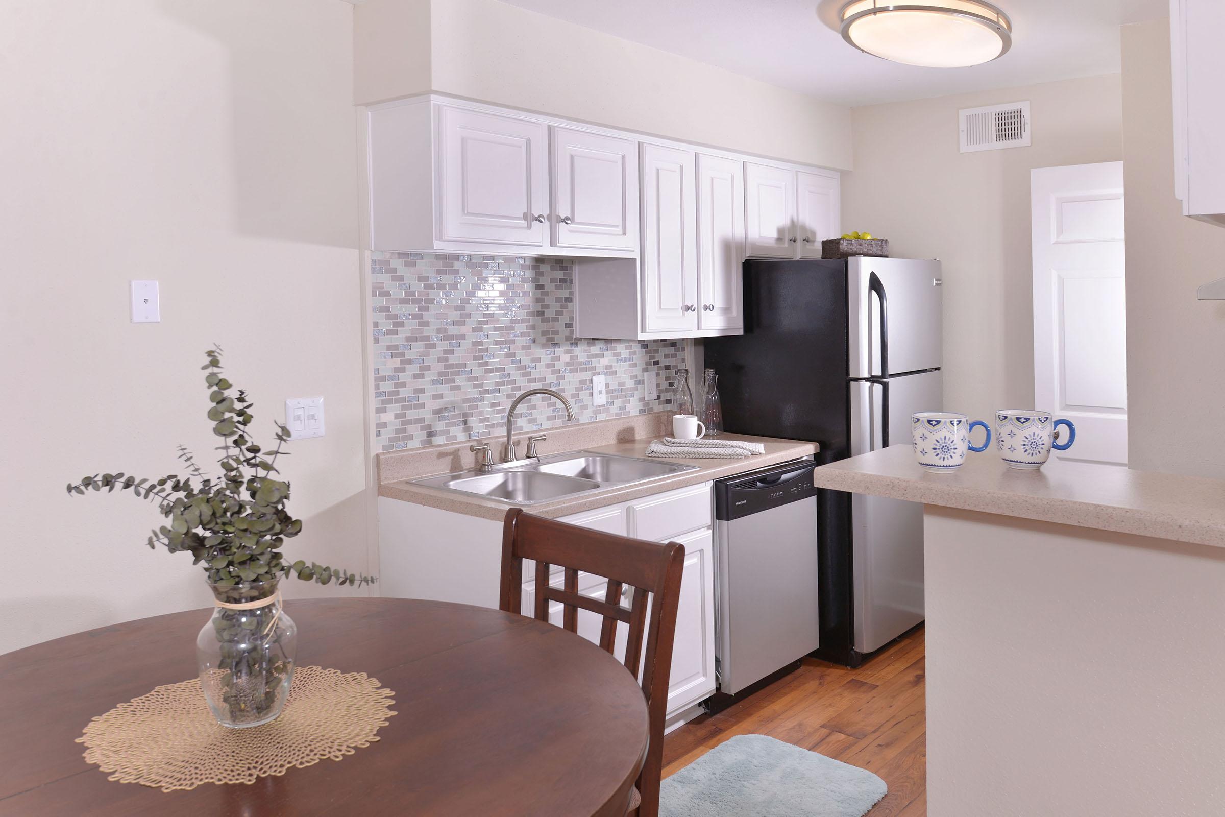 A clean and modern kitchen featuring white cabinetry, a mosaic tile backsplash, a stainless steel refrigerator, and a black dishwasher. There is a dining table with a lace placemat and a vase of greenery. The space has warm wooden flooring and soft lighting, creating a welcoming atmosphere.