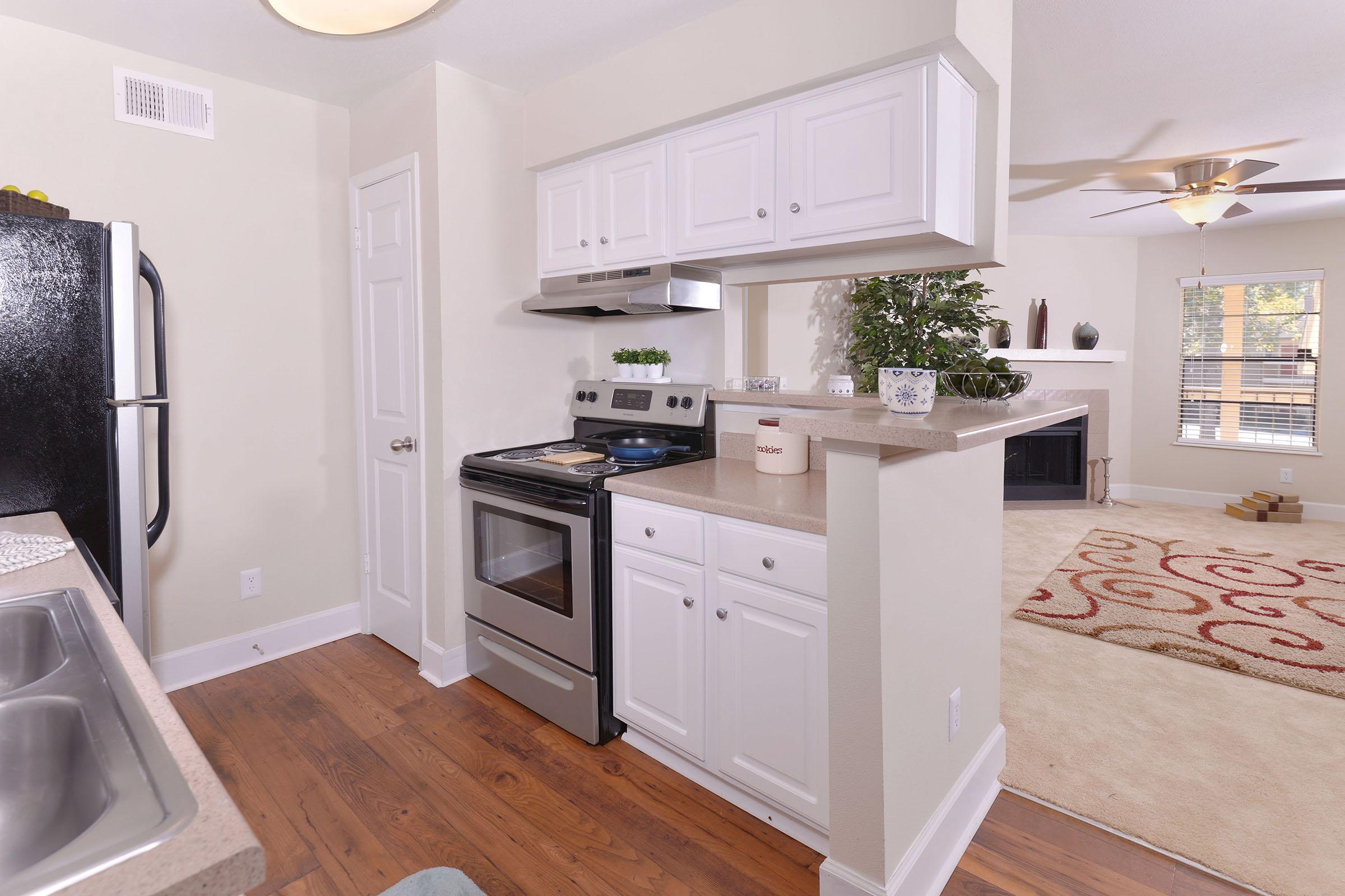 A modern kitchen featuring white cabinets, a stainless steel oven, and a black refrigerator. The countertop is decorated with plants, and there’s an adjacent living area with a cozy rug and a fireplace. Natural light illuminates the space through a nearby window.