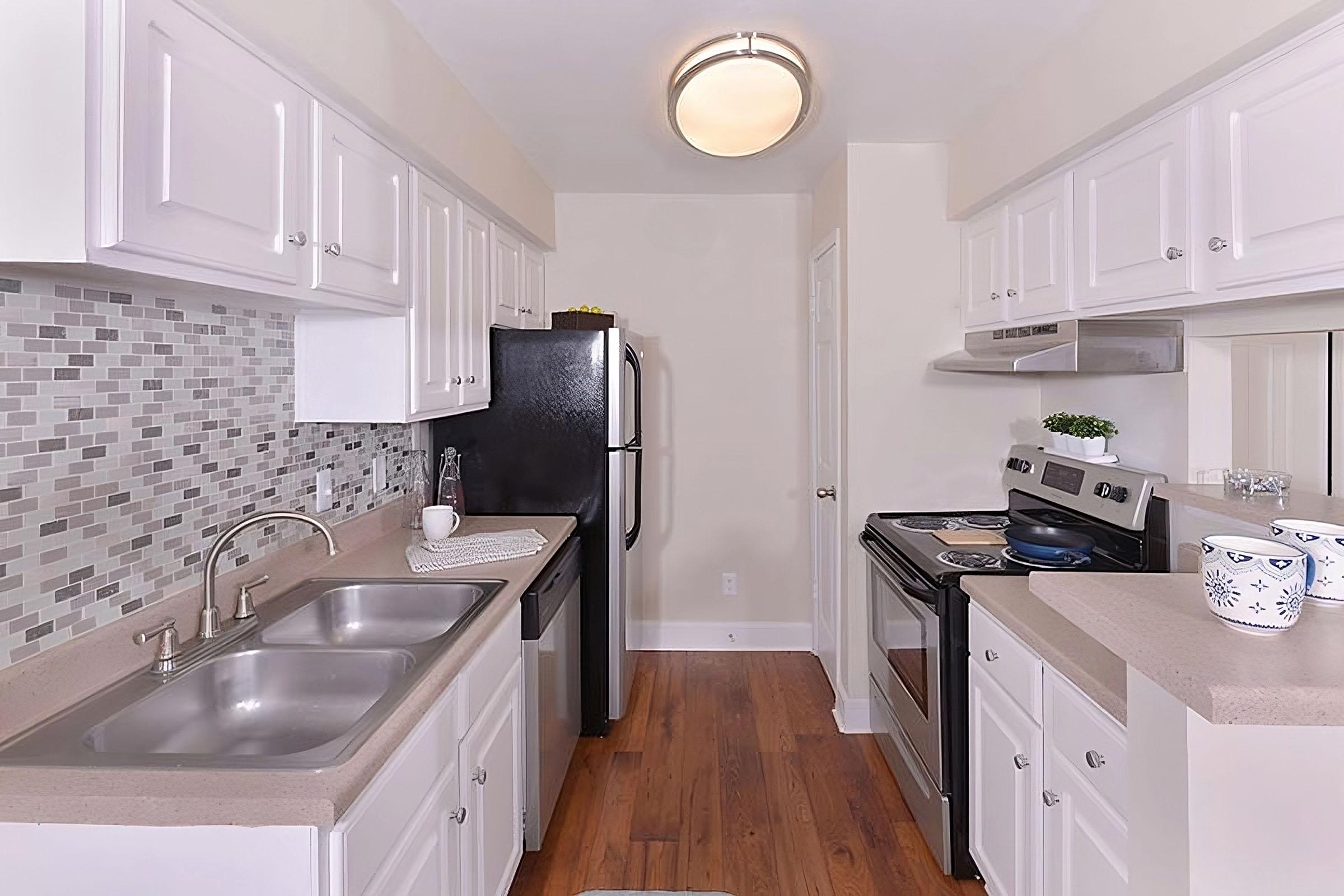 A modern kitchen featuring white cabinetry, a mosaic tile backsplash, stainless steel appliances, and a spacious countertop. The layout includes a double sink, a refrigerator, and an oven with a cooktop, set against warm wooden flooring, creating a bright and inviting atmosphere.