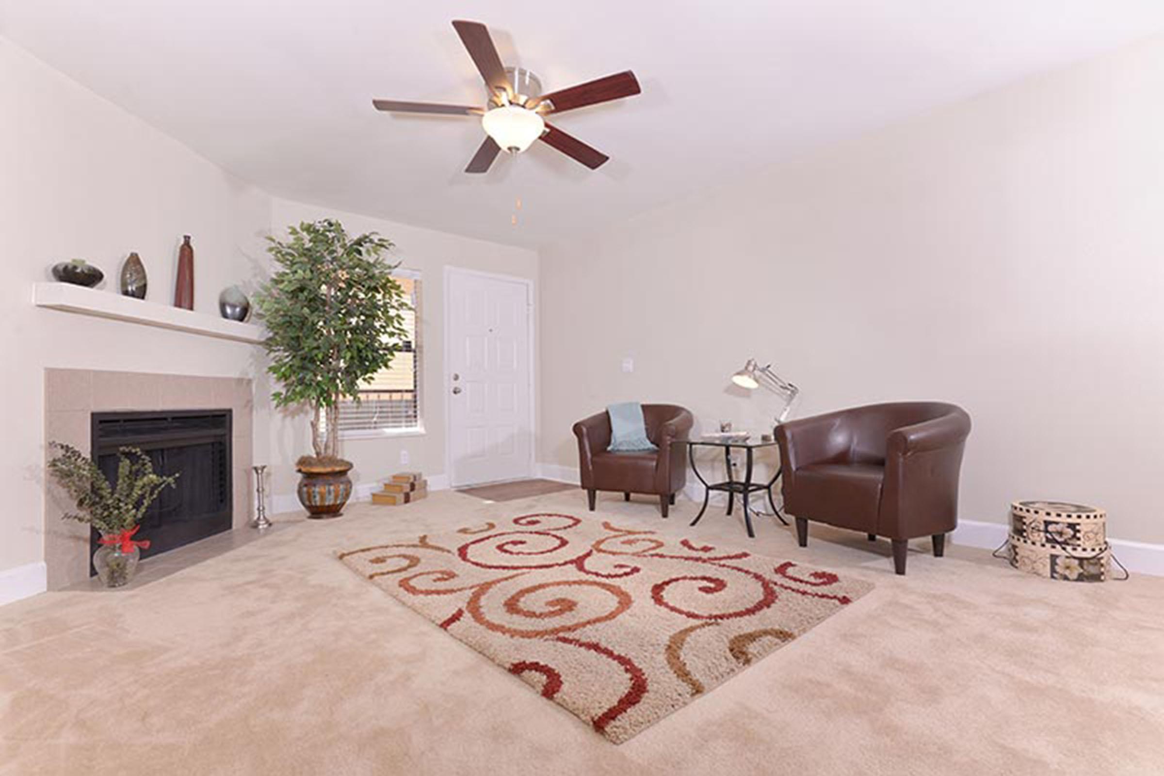 A cozy living room featuring two brown armchairs and a small glass table, with a decorative area rug displaying swirly patterns. A fireplace is set against a beige wall, and a potted plant adds a touch of greenery. The room is bright and uncluttered, creating a warm and inviting atmosphere.