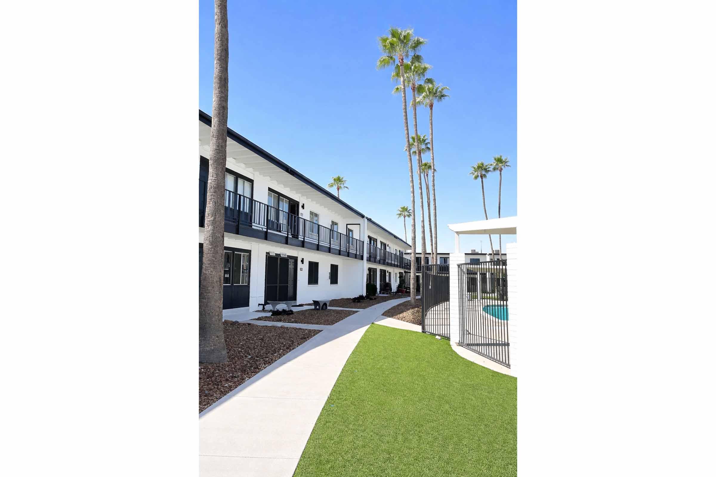A modern pathway leads past a white building with balconies, lined by tall palm trees against a clear blue sky. The scene features landscaped greenery and a pool area enclosed by fencing, creating a serene atmosphere typical of a sunny resort or apartment complex.