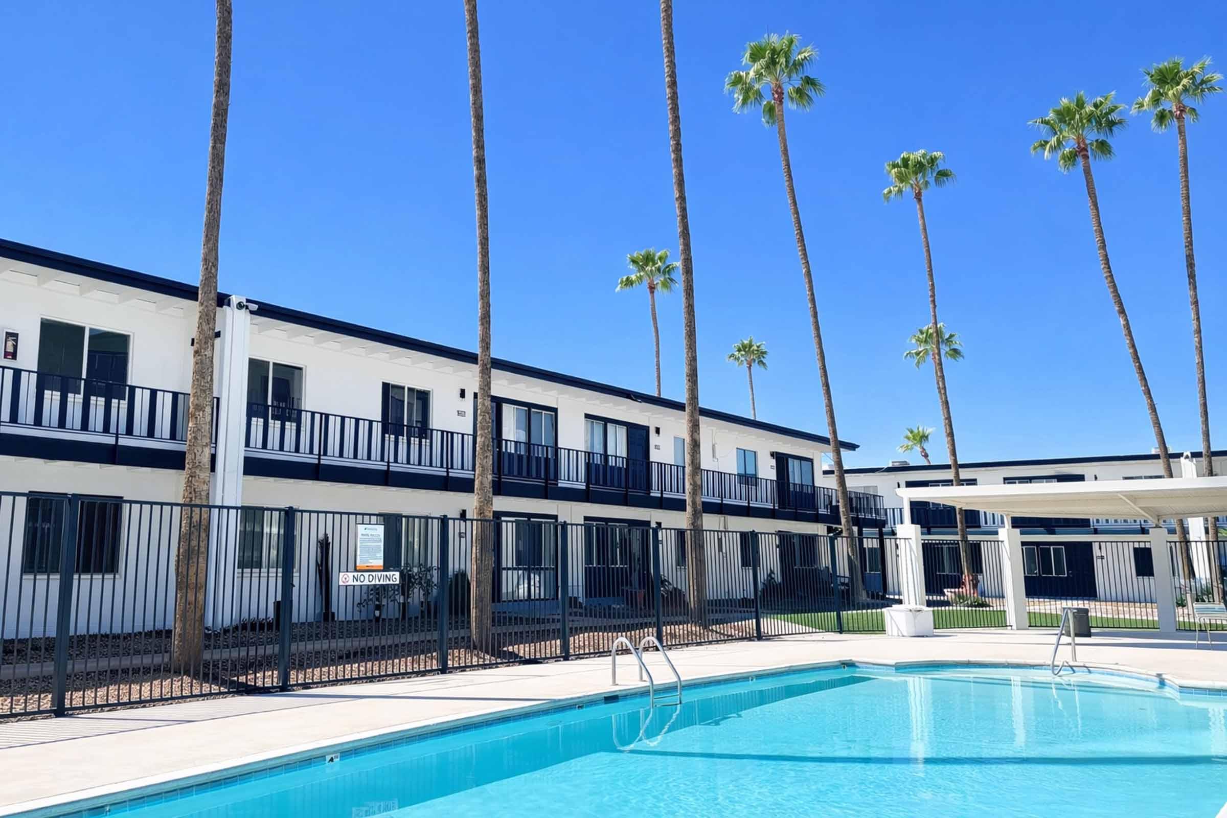 A bright blue sky over a swimming pool surrounded by a gated area. Palm trees line the backdrop of a two-story building with white walls and black railings. The scene conveys a relaxed, tropical atmosphere typical of a resort or hotel setting.