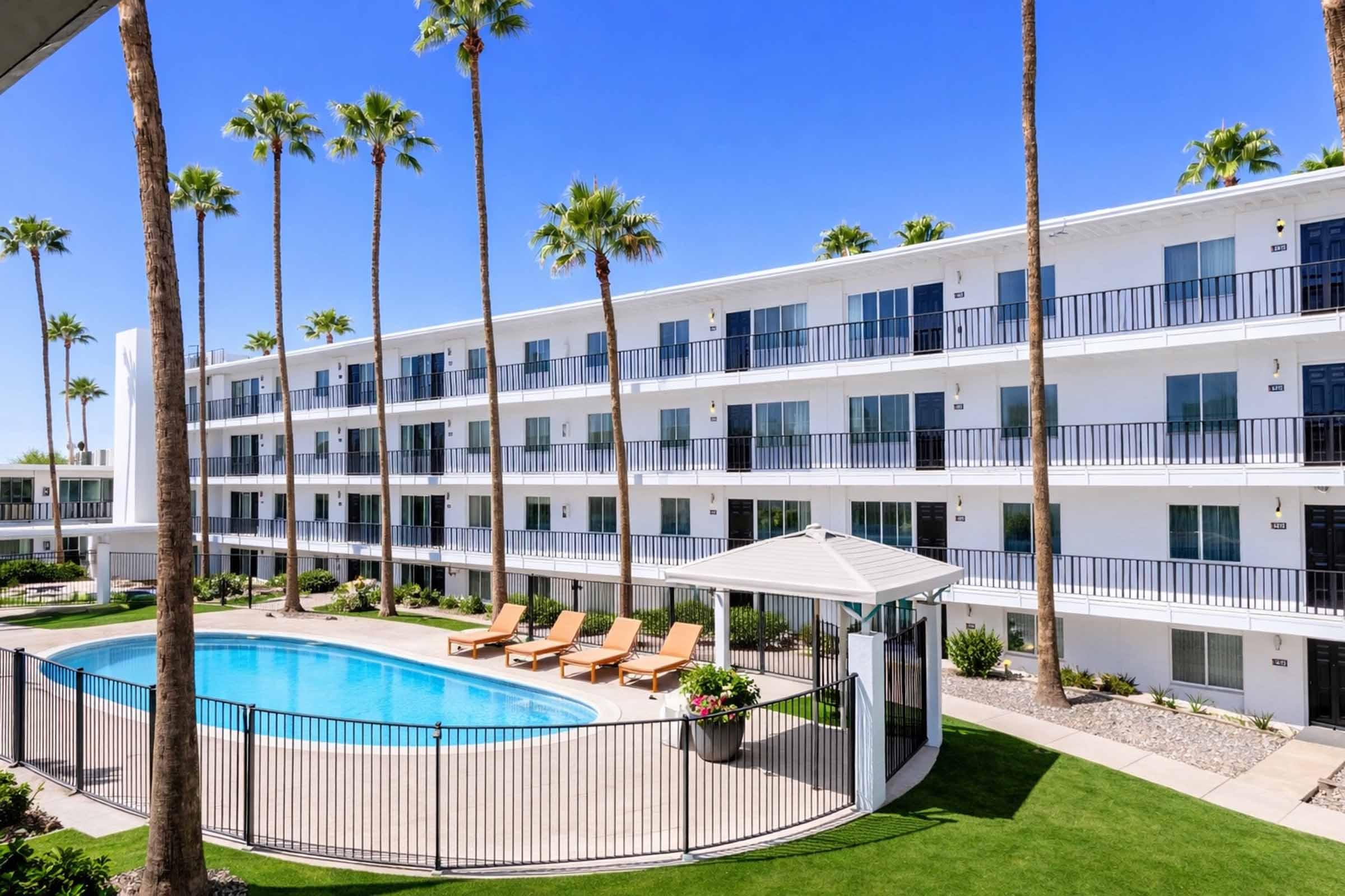Modern apartment complex featuring a rectangular swimming pool surrounded by palm trees. The building has multiple levels with balconies, and there are lounge chairs set up near the pool area. Clear blue sky enhances the bright, inviting atmosphere.