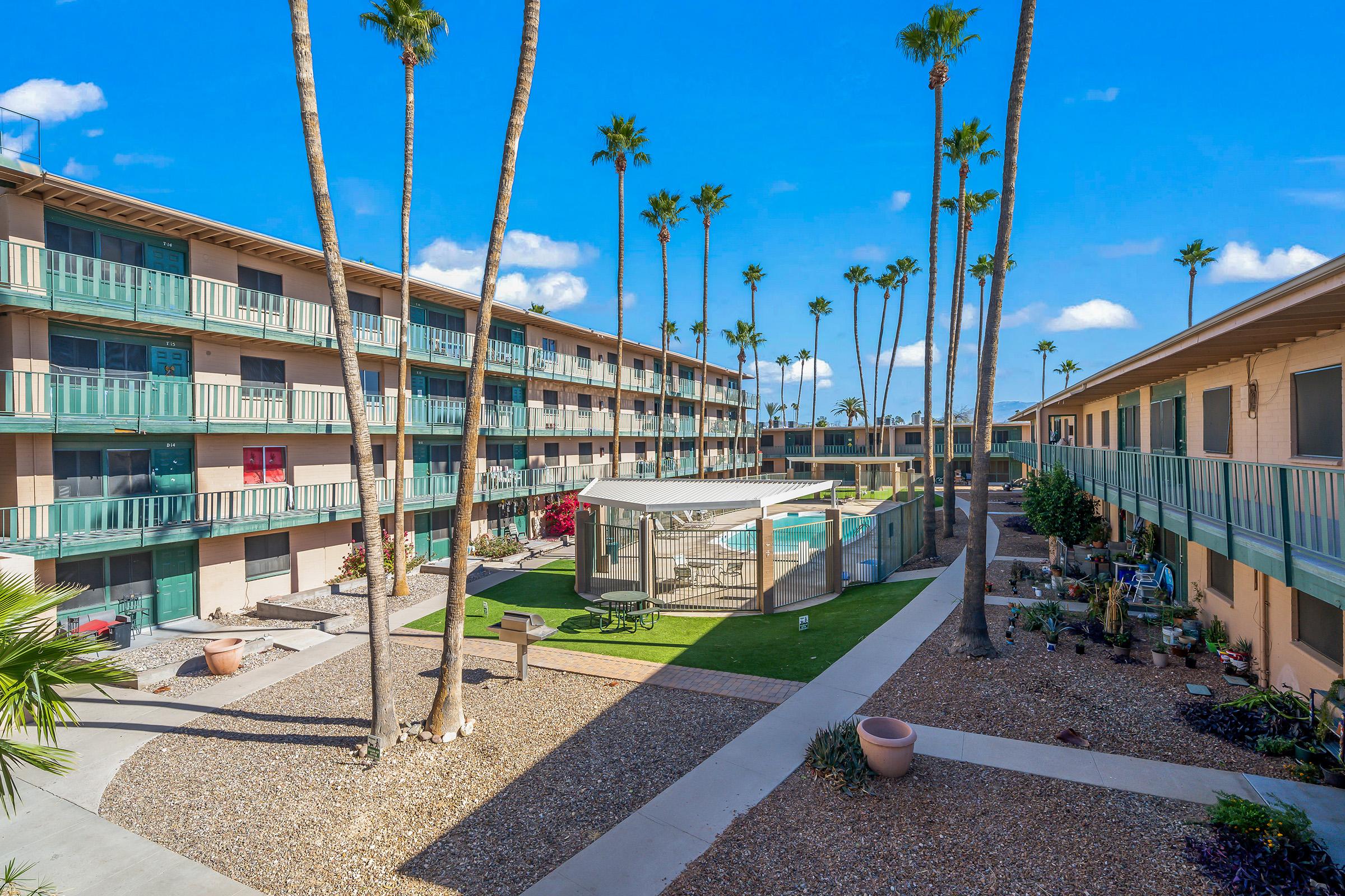 A view of a landscaped courtyard in an apartment complex featuring palm trees, a swimming pool surrounded by fencing, and two multi-story residential buildings. The setting is sunny with a clear blue sky, and there are pathways and decorative plants throughout the area.