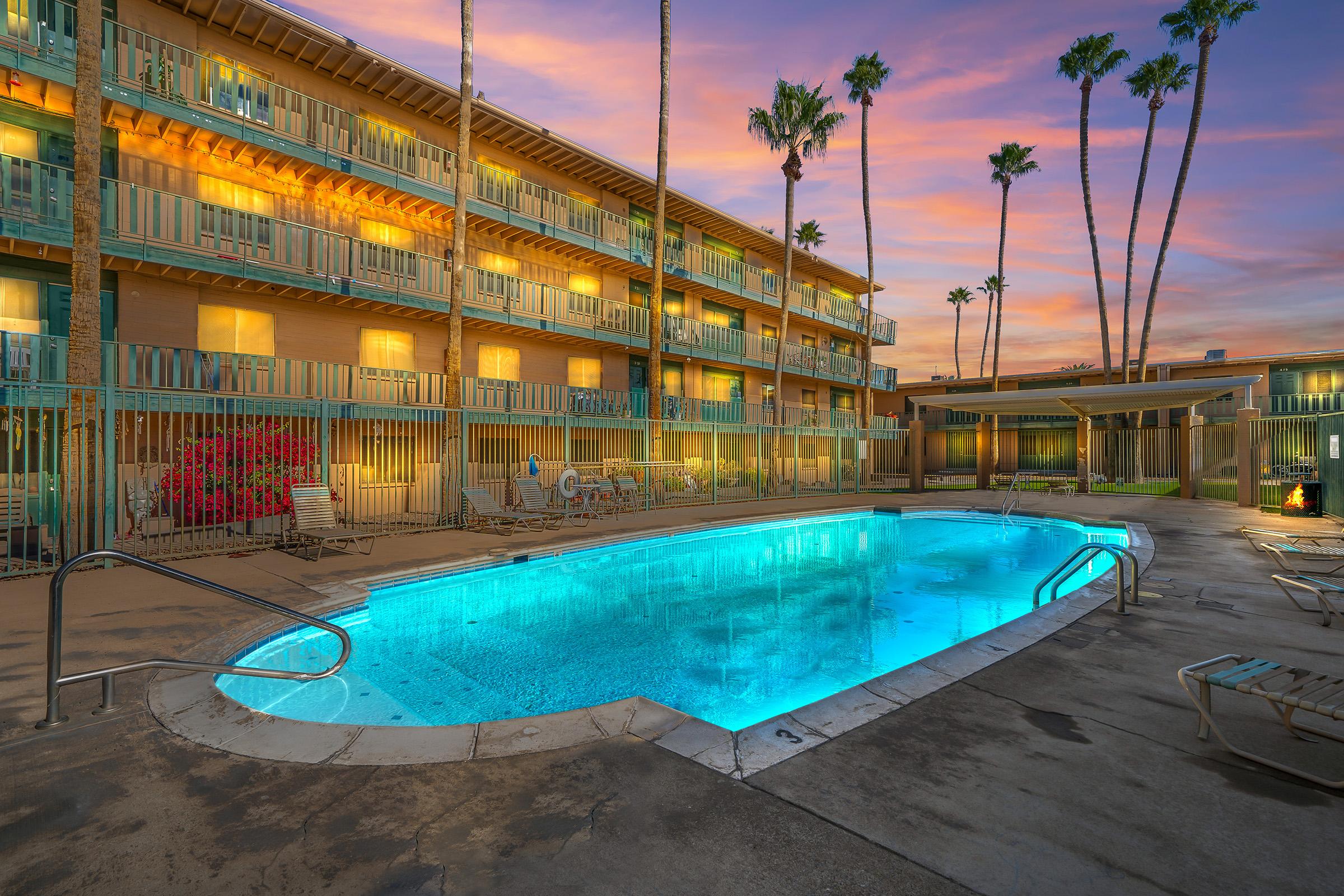 A serene outdoor pool area at dusk, surrounded by modern hotel balconies. Palm trees are silhouetted against a vibrant sunset sky with hues of orange and purple. Loungers are placed around the pool, creating a relaxing ambiance. The scene evokes a tranquil vacation atmosphere.