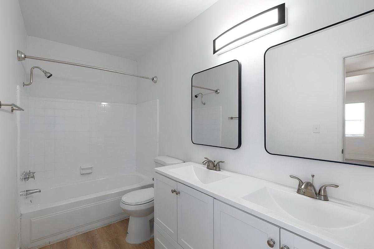 A modern bathroom featuring a bathtub with a shower, a dual-sink vanity with contemporary faucets, two large mirrors, and neutral-colored walls. The flooring is light wood, and there is a bright overhead light fixture. The space is minimalist and well-lit with natural light.