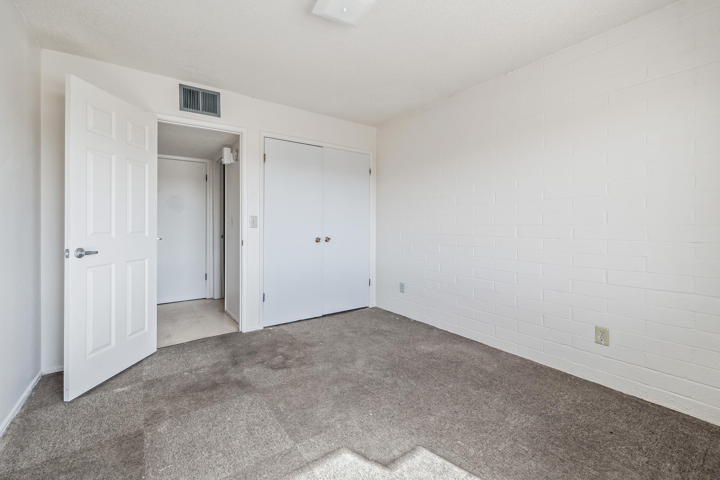 A bright, empty room with light-colored walls and a brown carpet. Two doors are visible: one leading to a closet and another to a hallway. Natural light enters from a window, creating an open and spacious feel. The room has minimal furnishings, emphasizing its versatility for various uses.