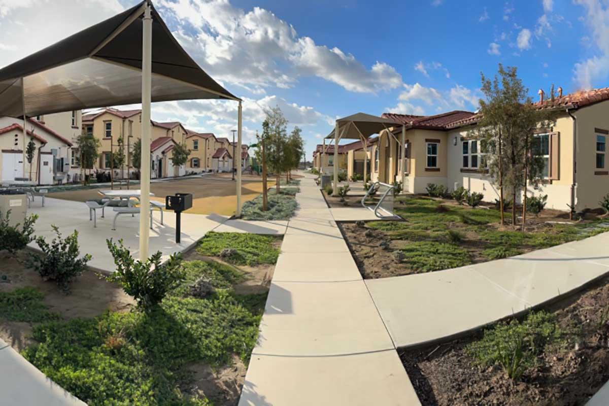 A wide view of a residential area featuring modern houses, a paved walkway, and shaded seating areas.