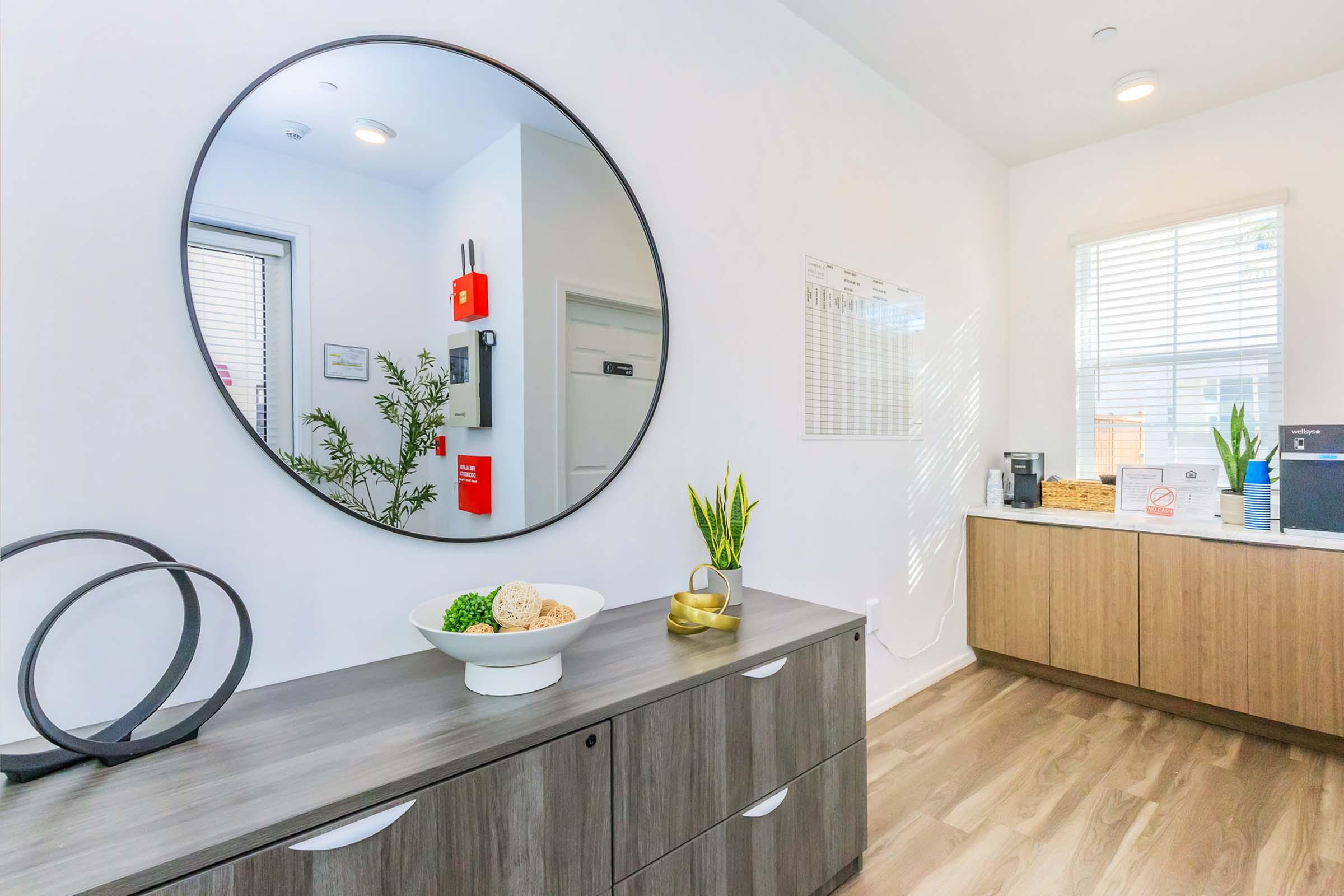 A bright, modern interior featuring a circular mirror on a white wall. A wooden cabinet holds decorative plants and a bowl with items, while a coffee machine and calendar are seen on a nearby countertop. Natural light flows in through a window, enhancing the contemporary feel of the space.