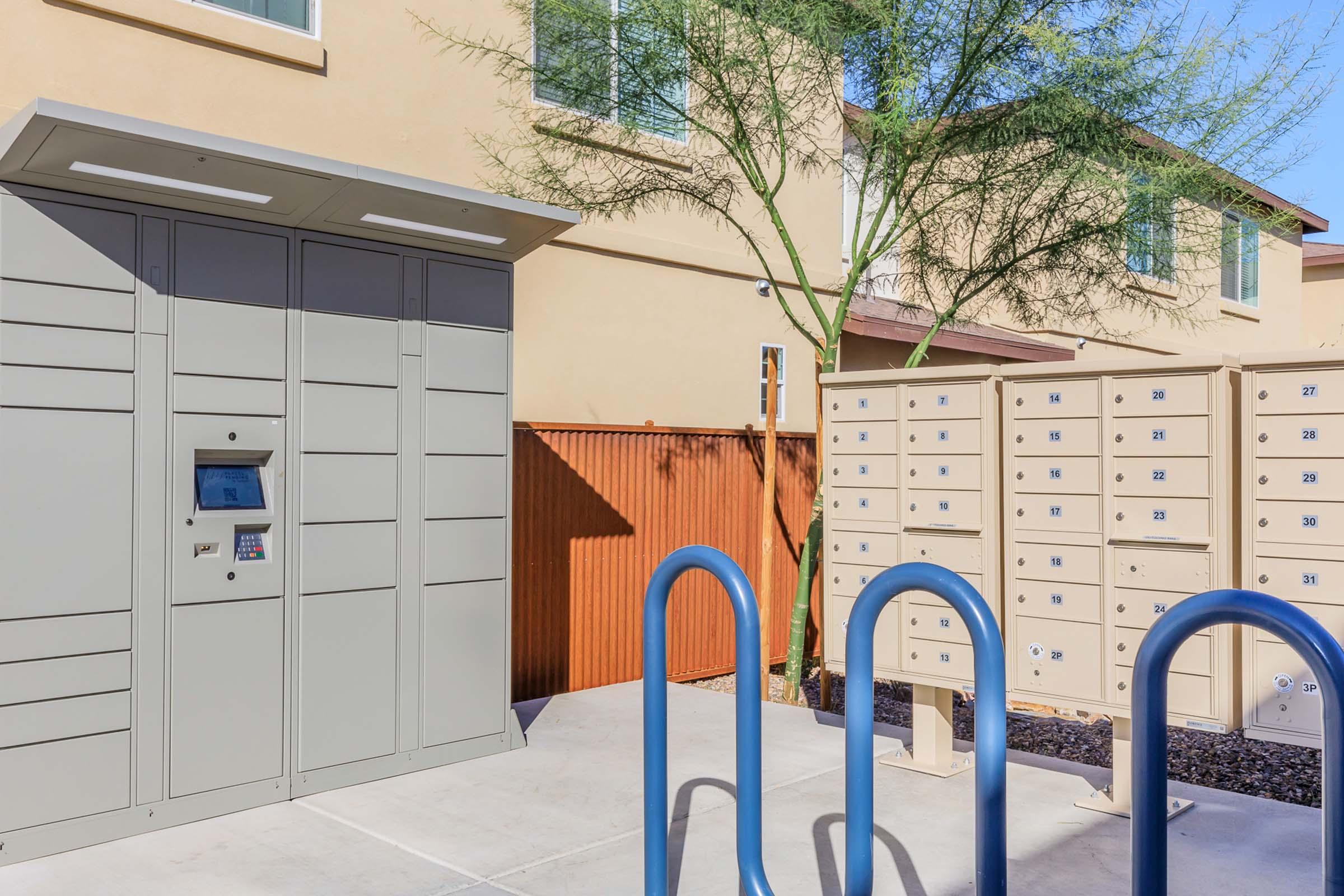 A modern package delivery kiosk in an outdoor area with various mailboxes. The kiosk features a digital screen and buttons for user interaction, while the mailboxes are lined up on the right. There are blue bike racks in the foreground, and a tree is visible in the background against a residential building.