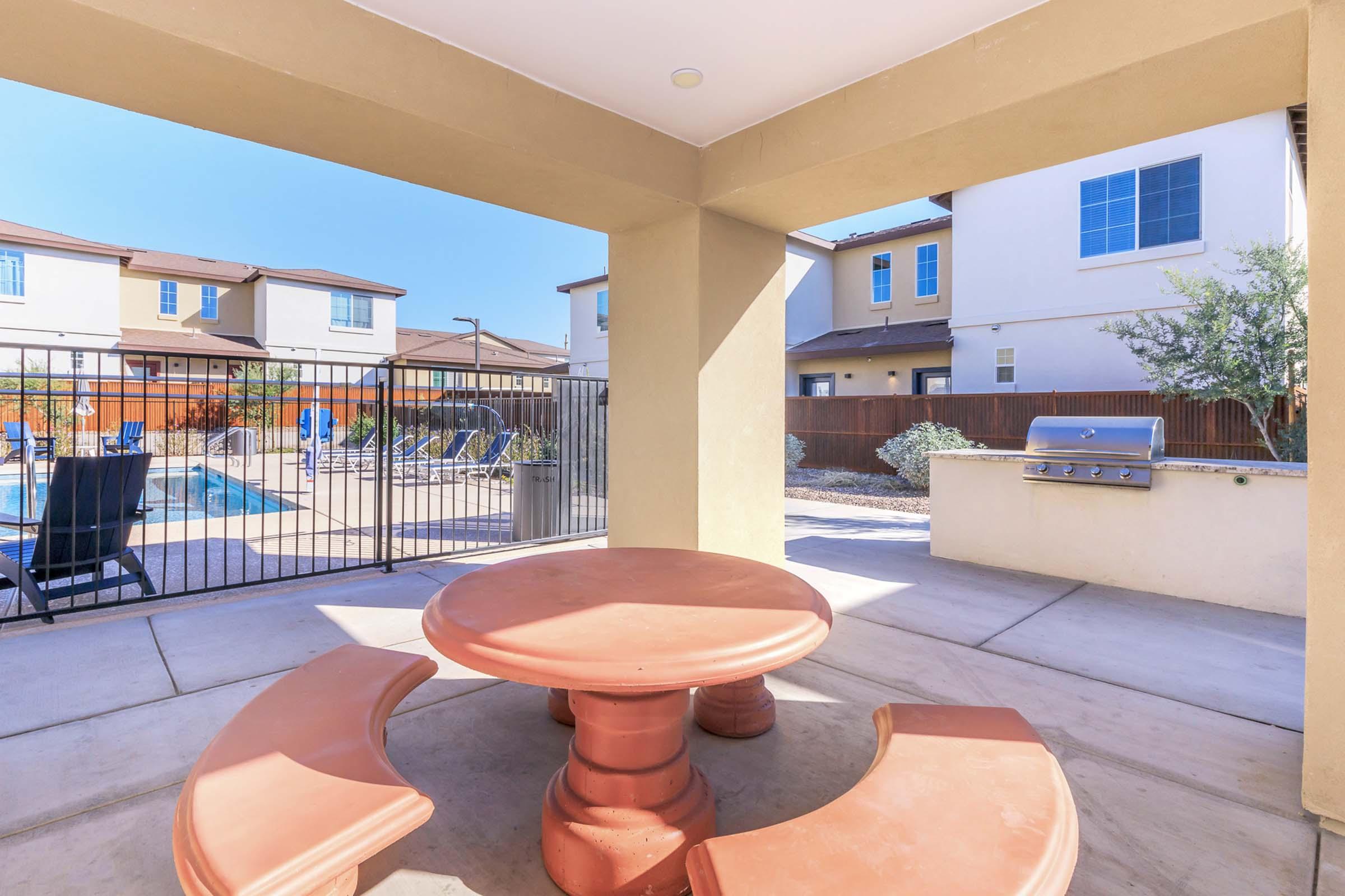 Outdoor seating area with a circular stone table and bench, overlooking a pool area surrounded by lounge chairs. In the background, there are neighboring houses and a barbecue grill, all under clear blue skies. The space is well-lit and designed for relaxation and social gatherings.