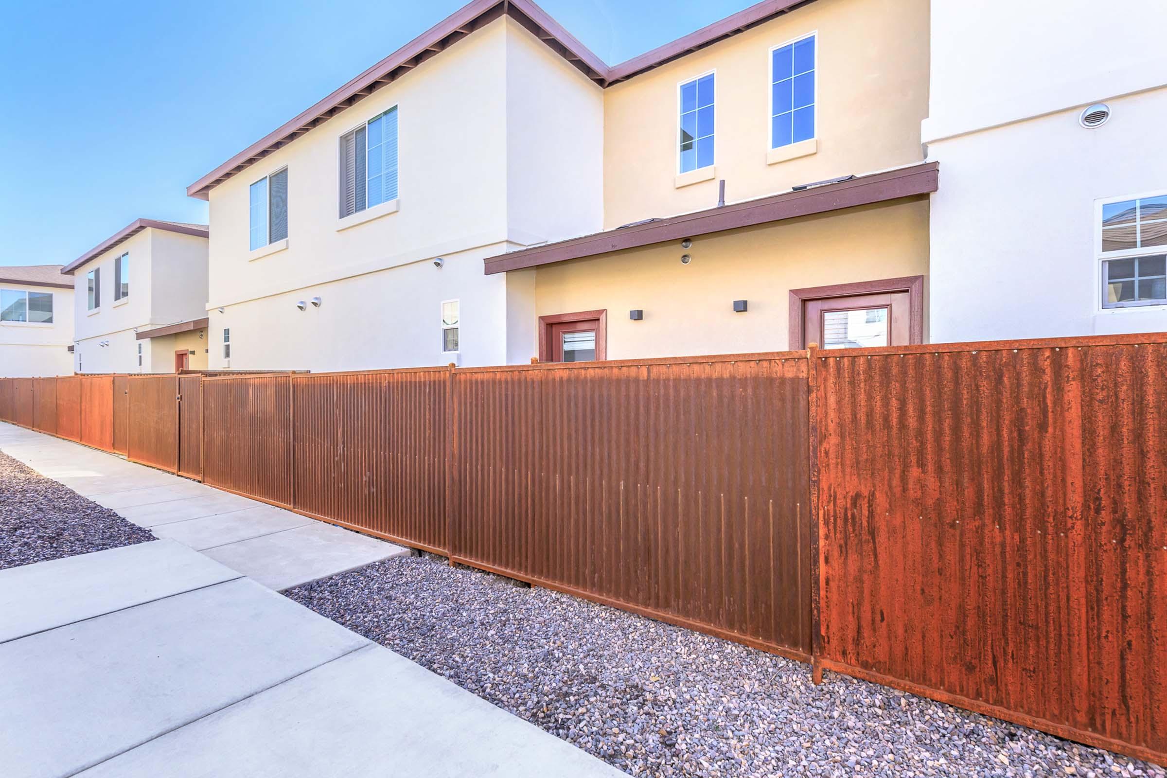A view of a residential area featuring two modern townhouse buildings with concrete walkways. A rust-colored corrugated metal fence runs along the side, with small gravel landscaping visible. The buildings have large windows and are painted in neutral colors, creating a contemporary look.