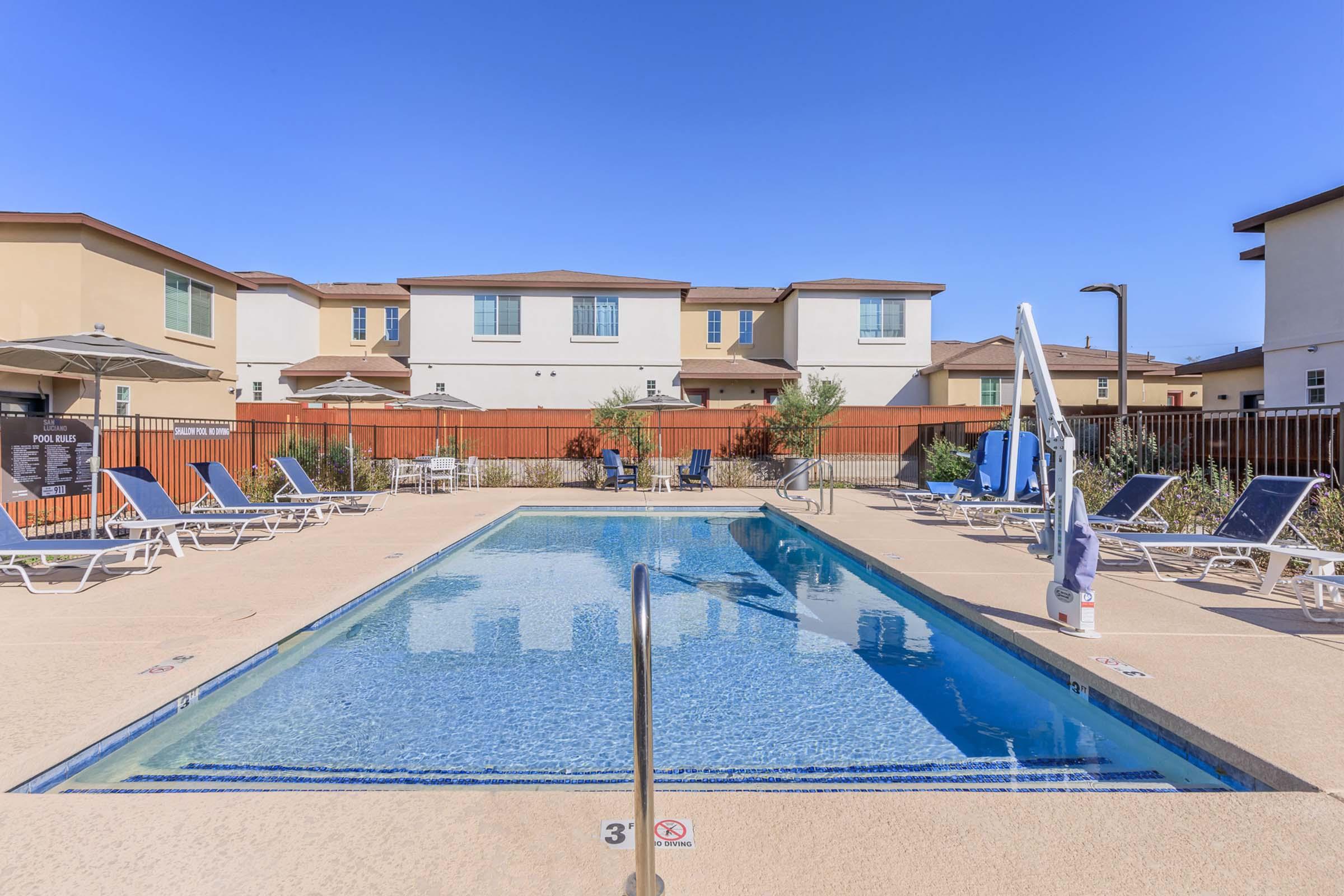 A clear swimming pool surrounded by lounge chairs on a sunny day. In the background, there are residential buildings with multiple stories and a wooden fence enclosing the pool area. The sky is blue with no clouds.