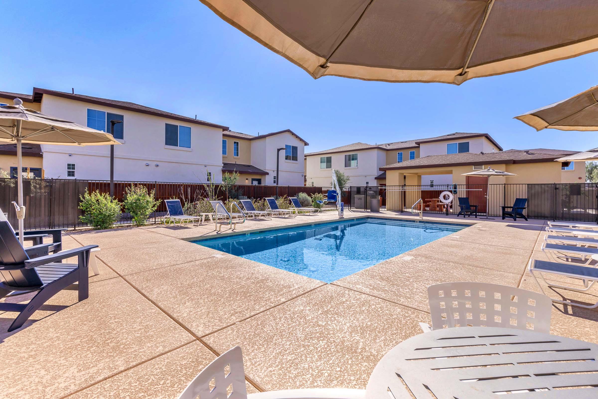 A clear swimming pool surrounded by lounge chairs and umbrellas, with residential buildings in the background. The area is sunny and well-maintained, offering a relaxing outdoor space.