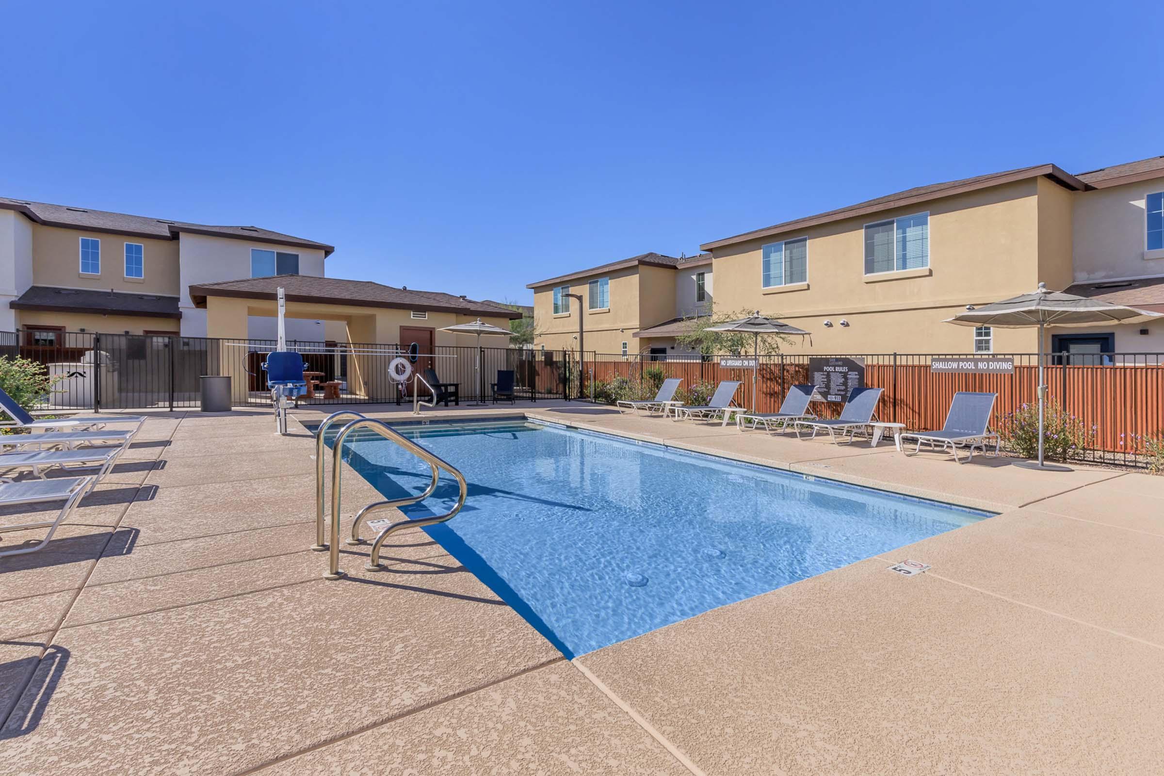 A clear blue swimming pool surrounded by a patio with lounge chairs. There are umbrellas for shade and a fence enclosing the area. In the background, there are contemporary two-story buildings under a bright blue sky.