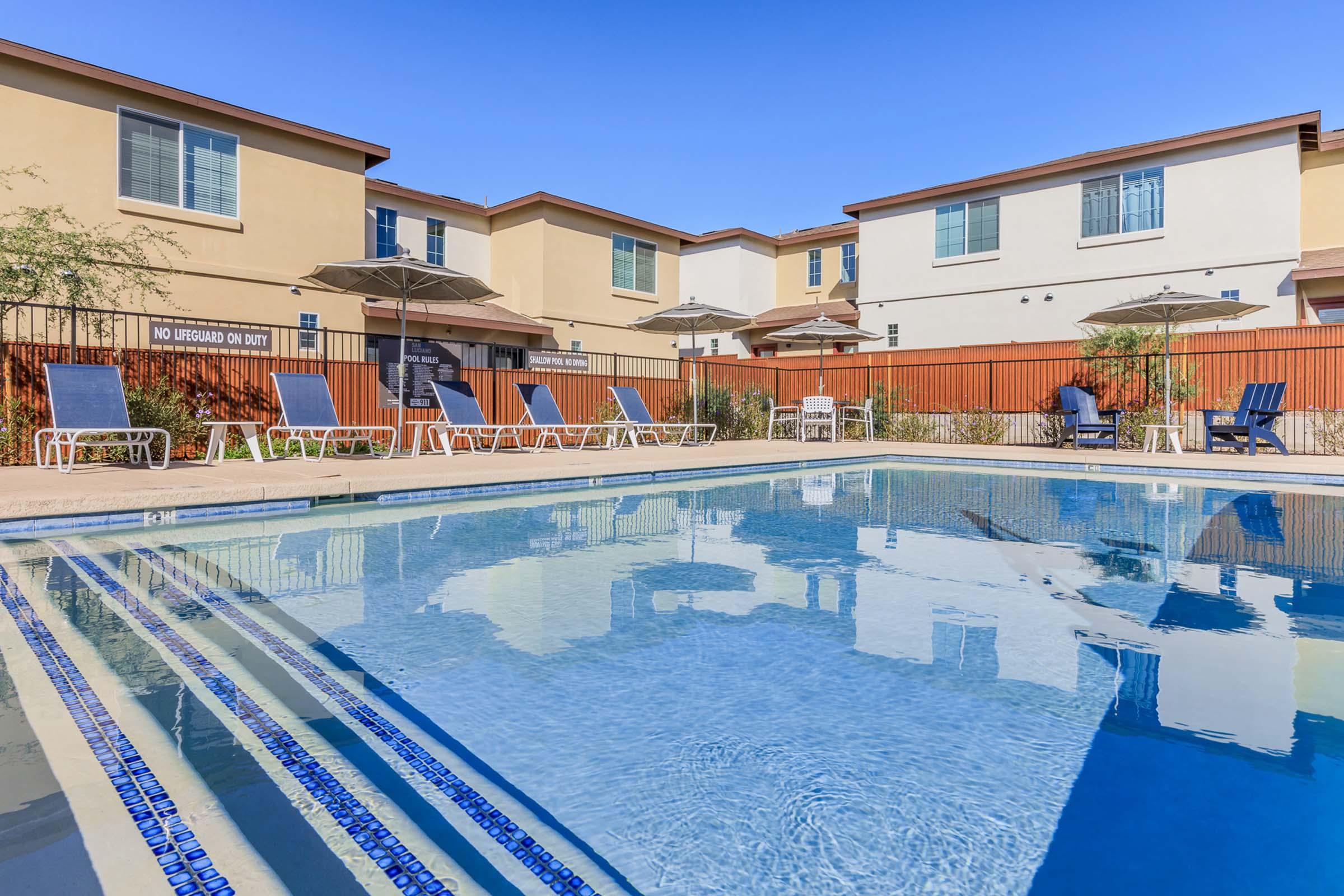 A clear swimming pool surrounded by lounge chairs and umbrellas, with residential buildings in the background. The sky is bright blue, and the pool area is well-maintained, creating a relaxing atmosphere. A sign indicates "No Lifeguard on Duty" near the pool.
