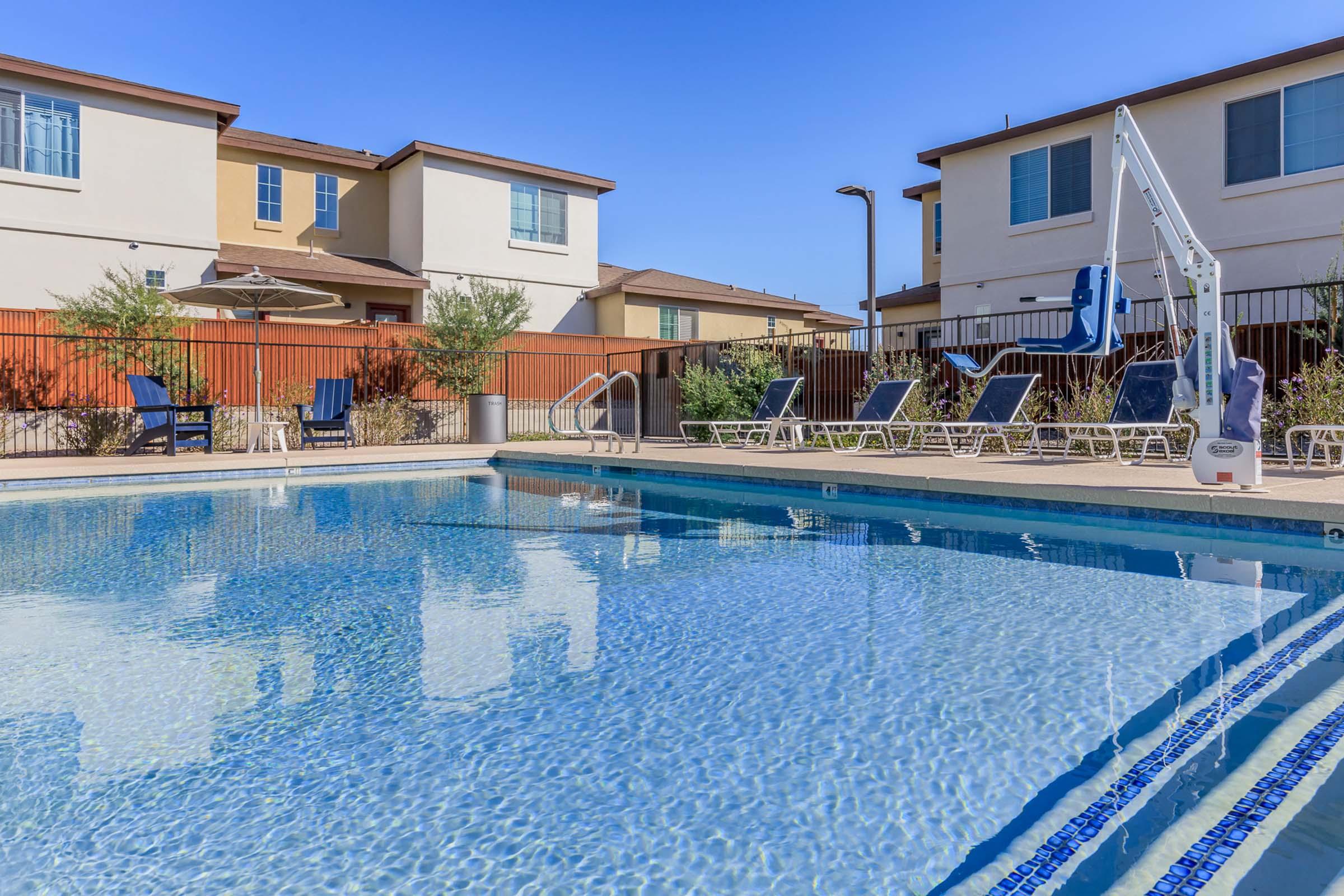 A clear swimming pool surrounded by lounge chairs and greenery. In the background, there are residential buildings with blue skies overhead. The area is well-maintained, inviting relaxation and leisure. A shaded seating area with an umbrella is also visible.