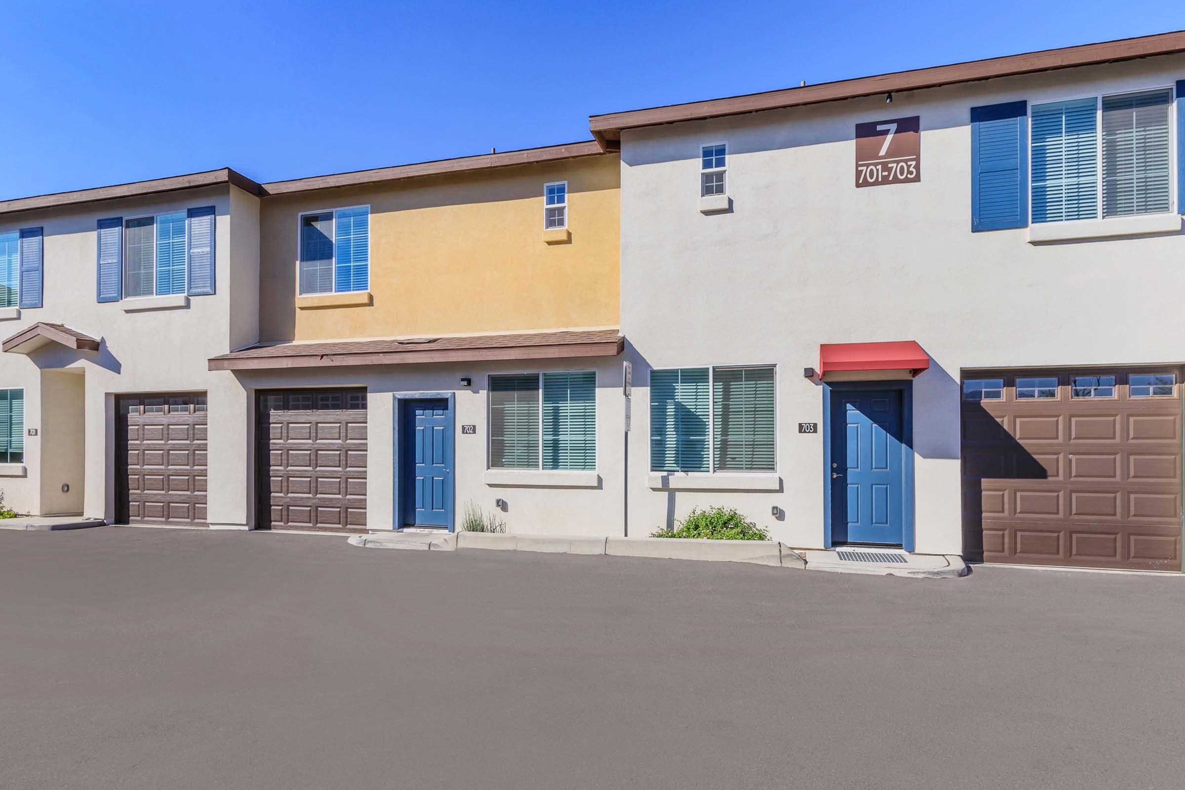 Row of modern townhouses with two garages, one blue door, and light yellow and white facades. Each unit features multiple windows with blue trim. The sky is clear and blue, and the asphalt driveway is well-maintained. A sign reads "7 701-703" above the entrance.