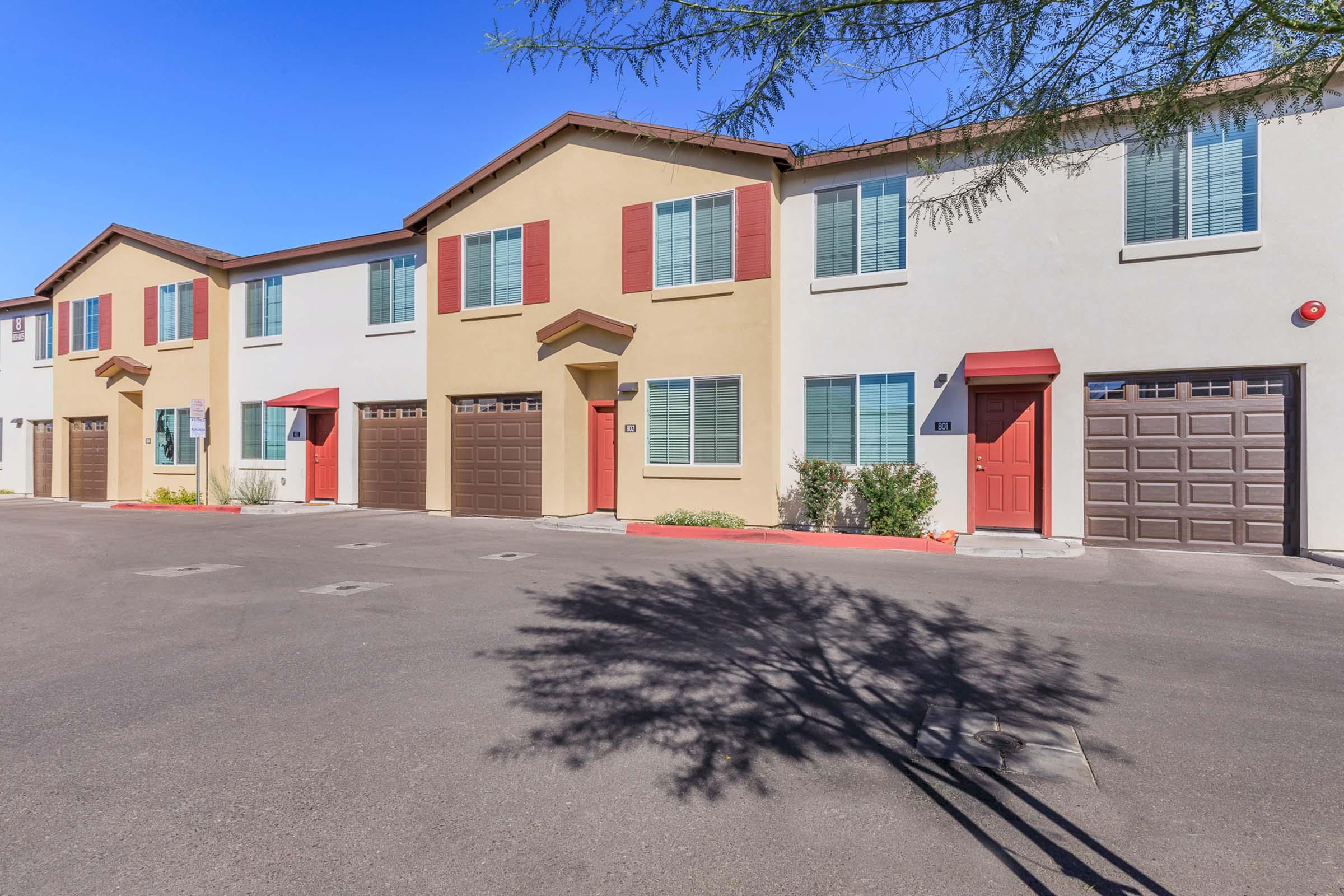 A row of modern townhouses featuring beige and cream exteriors with red trim. Each unit has a garage door and various windows, casting shadows on the pavement. The scene is set in a clear blue sky, surrounded by desert landscaping.