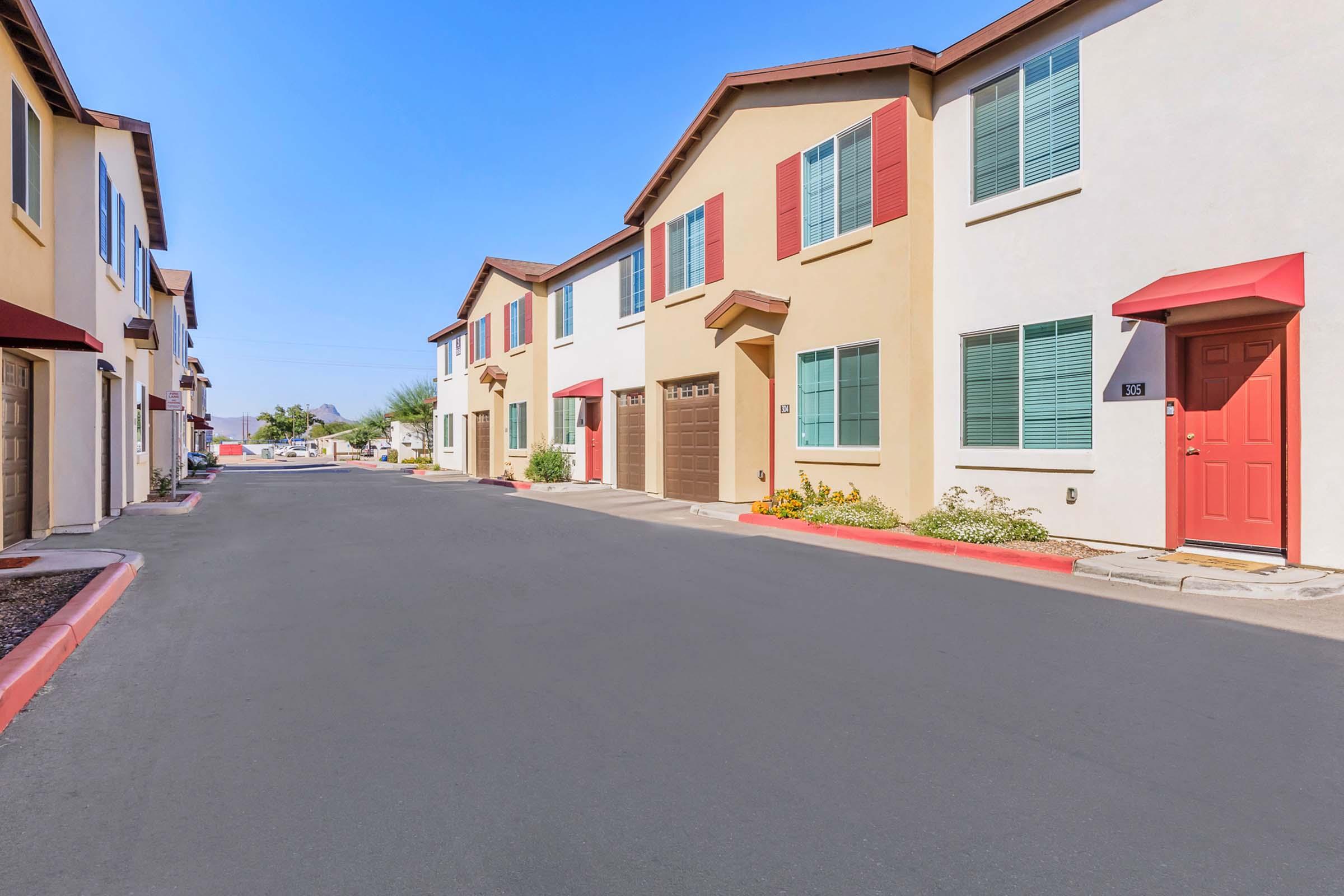 A row of modern two-story townhouses with colorful facades and red doors. The street is clean with a clear blue sky overhead. Small flower beds and shrubs are visible in front of some units, adding greenery to the scene.