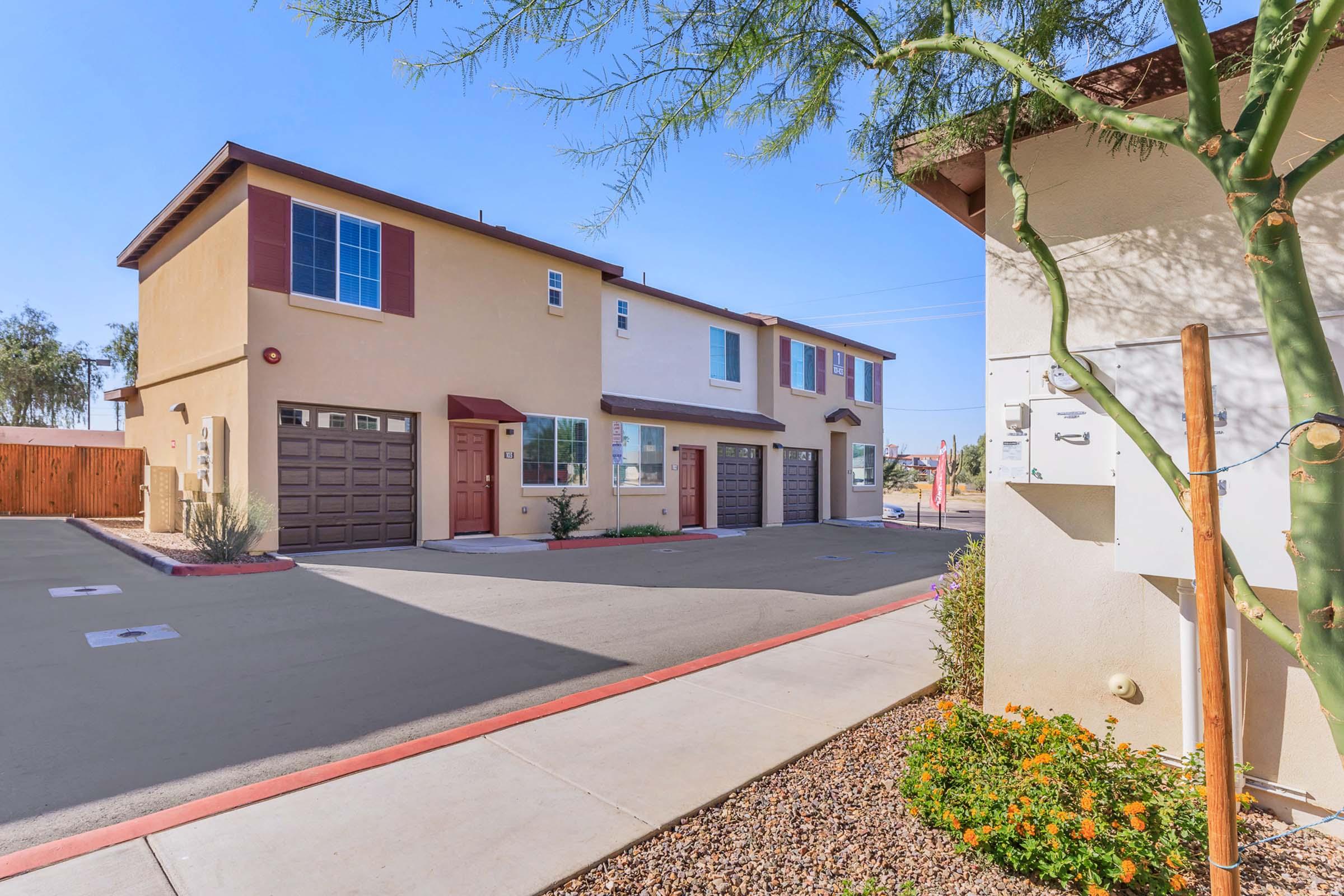 A modern residential building with two stories, featuring beige and brown exteriors. The property includes a paved parking area with garages, landscaped with orange flowers and surrounding desert vegetation. Clear blue skies are visible above, indicating a sunny day.