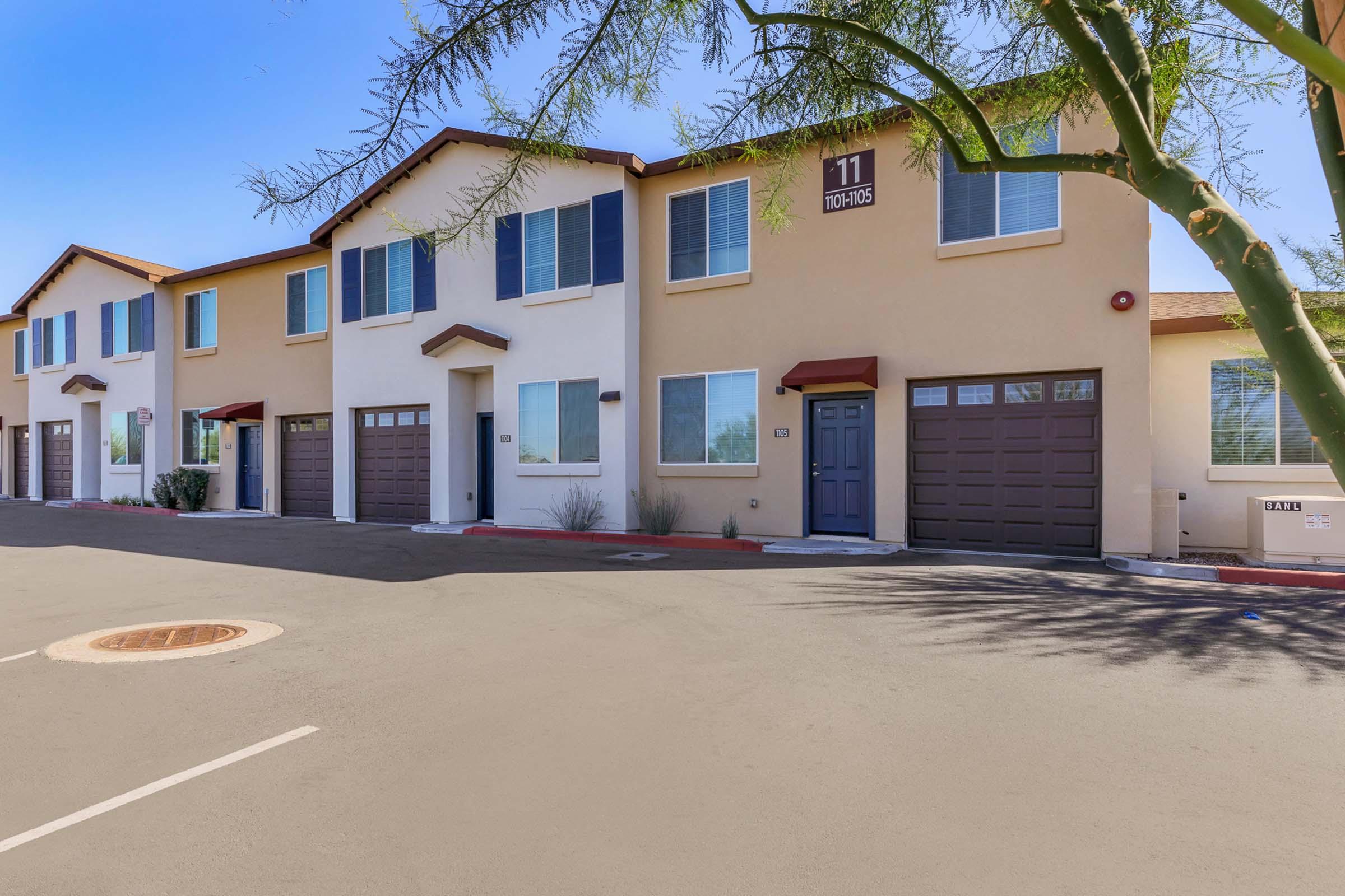 Row of townhouses with beige and brown exteriors. Each unit features a garage and windows with blue shutters. The street is paved with parking spaces visible. A tree is partially in the frame on the right side, providing some shade. The overall scene is bright and sunny, suggesting a residential neighborhood.