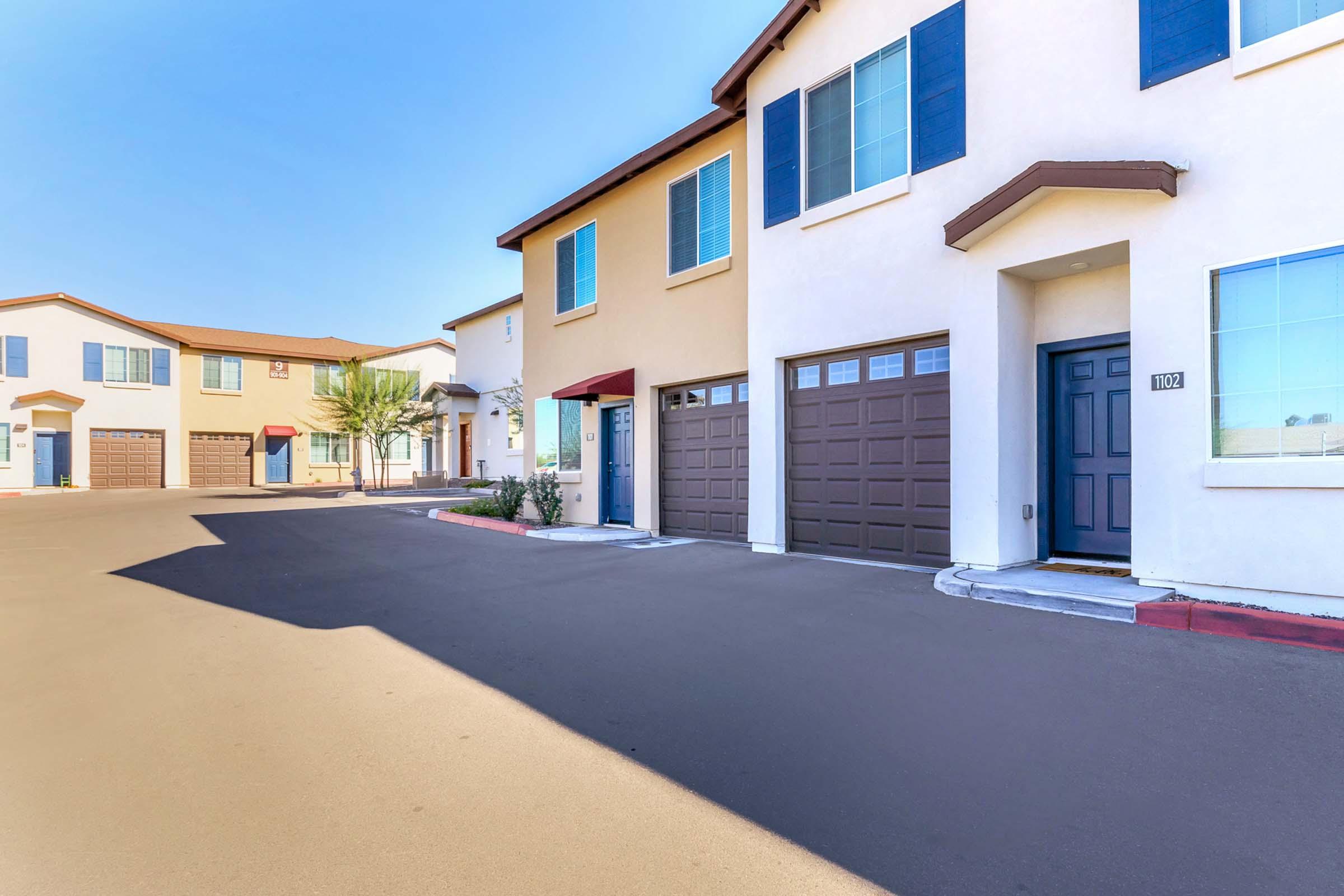 Row of modern two-story townhouses in a residential neighborhood. The buildings feature light-colored exteriors with dark shutters and single-car garages. A clear blue sky is visible overhead, and the pavement appears freshly sealed, indicating a well-maintained environment.