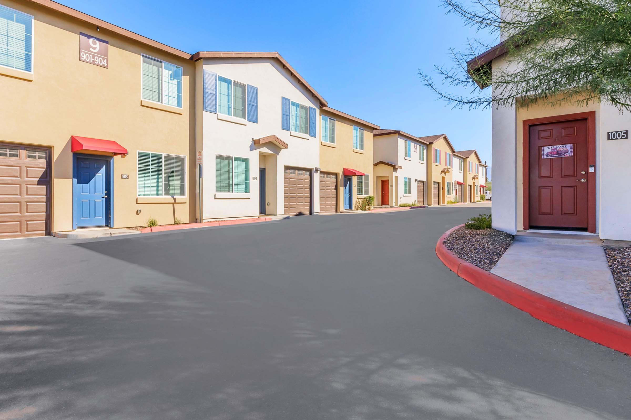 A quiet residential street lined with colorful townhouses featuring red awnings over the doors. The houses have various shades of beige and brown exteriors, with a clear blue sky overhead. A smooth asphalt road curves gently, leading to additional homes in the distance.