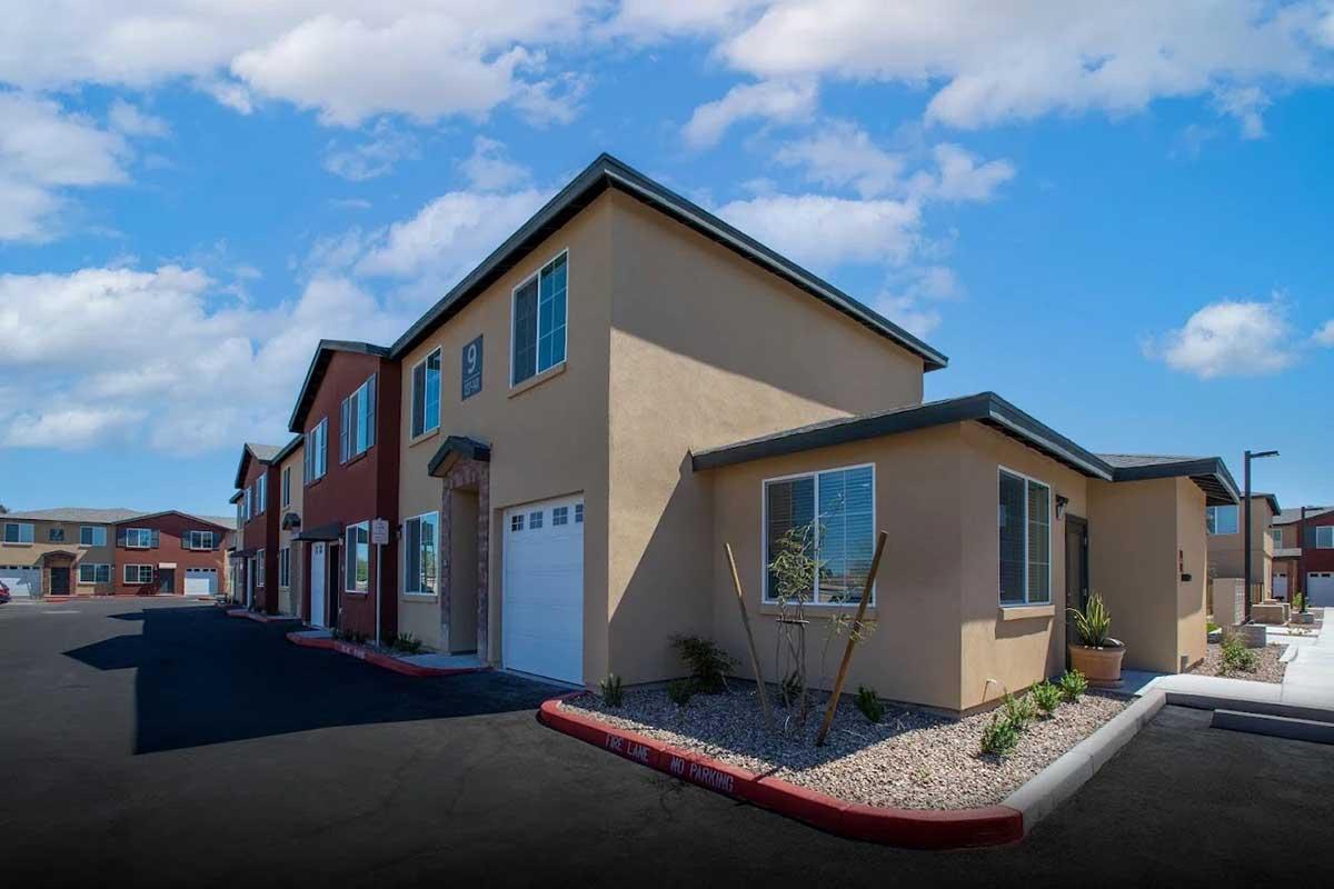 A modern residential building with a blend of tan and red exterior colors, featuring multiple windows and a garage. The foreground has a well-maintained parking area, while the background shows additional similar buildings under a bright blue sky with scattered clouds.