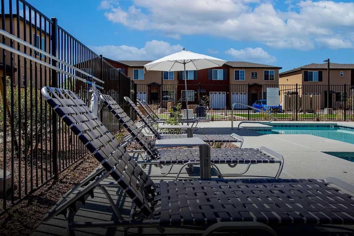 A pool area featuring several lounge chairs, a large umbrella, and a clear sky with fluffy clouds. In the background, there are several residential buildings. The pool is surrounded by a black fence, creating a relaxing outdoor space for leisure.