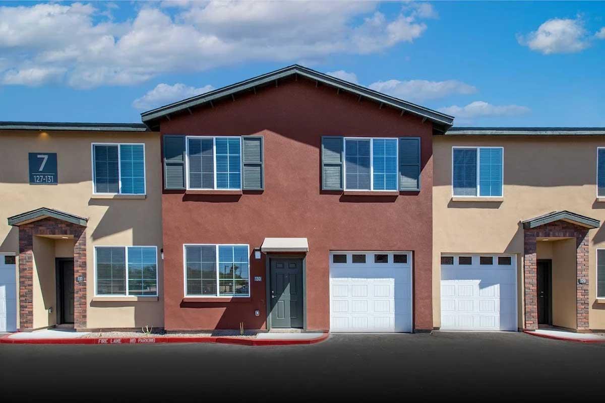 A modern apartment building with two stories and several units. The facade features a mix of cream and reddish-brown colors, with large windows and garages. The area is paved and shows a clear blue sky with fluffy clouds.