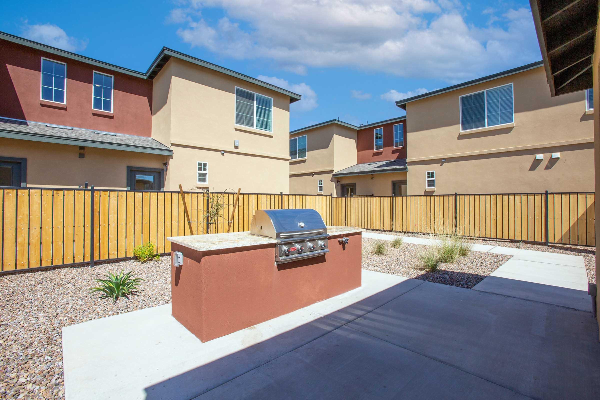 A well-maintained outdoor space featuring a barbecue grill on a concrete patio, surrounded by gravel and low-maintenance landscaping. The area is enclosed by a wooden fence, with modern multi-story buildings in the background under a clear blue sky.