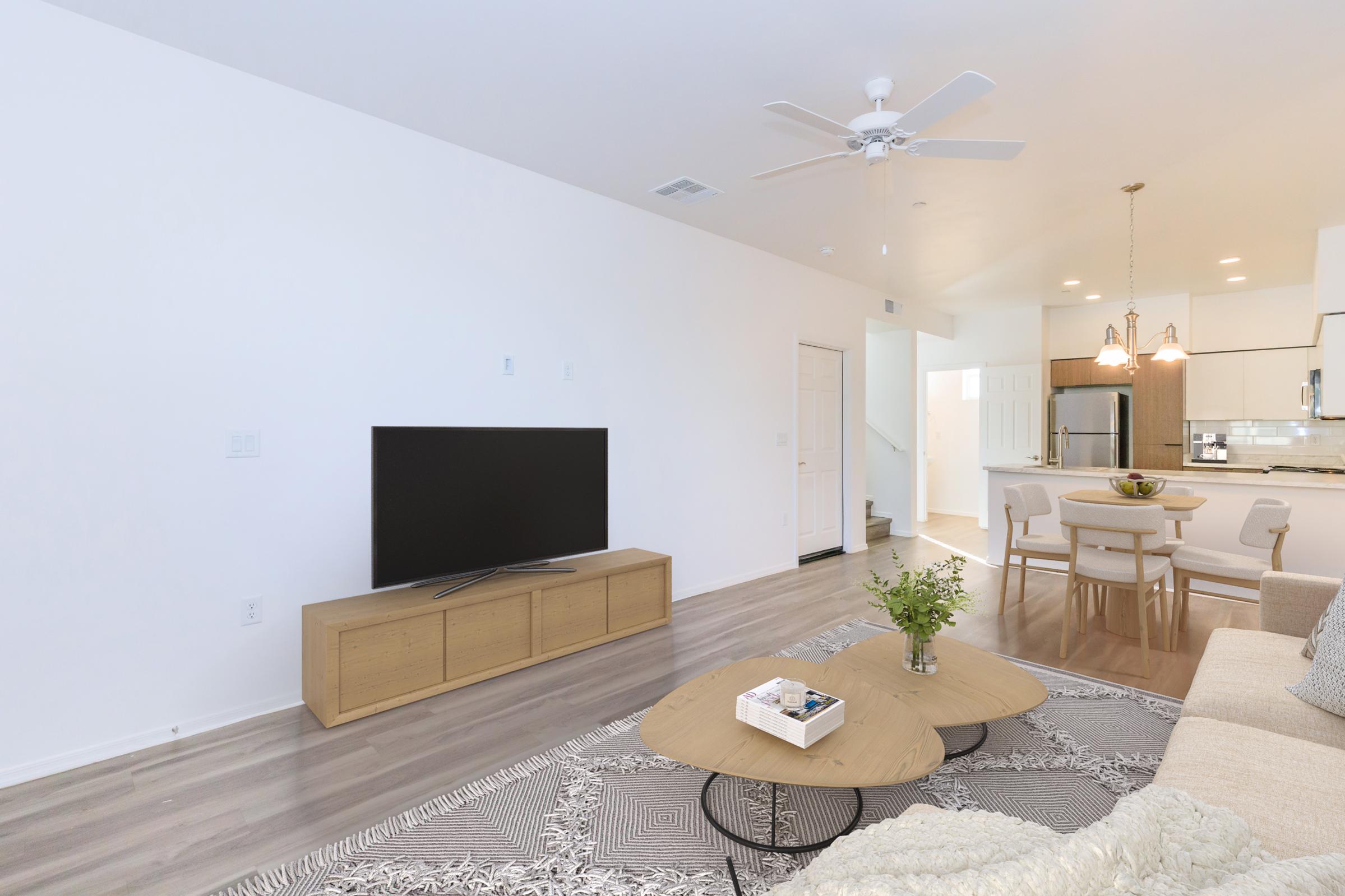 Modern living room featuring a large flat-screen TV on a wooden console, a stylish coffee table, and a beige sofa with textured throw. Light hardwood flooring and a white wall create a spacious atmosphere. In the background, a dining area with a round table and a staircase is visible.
