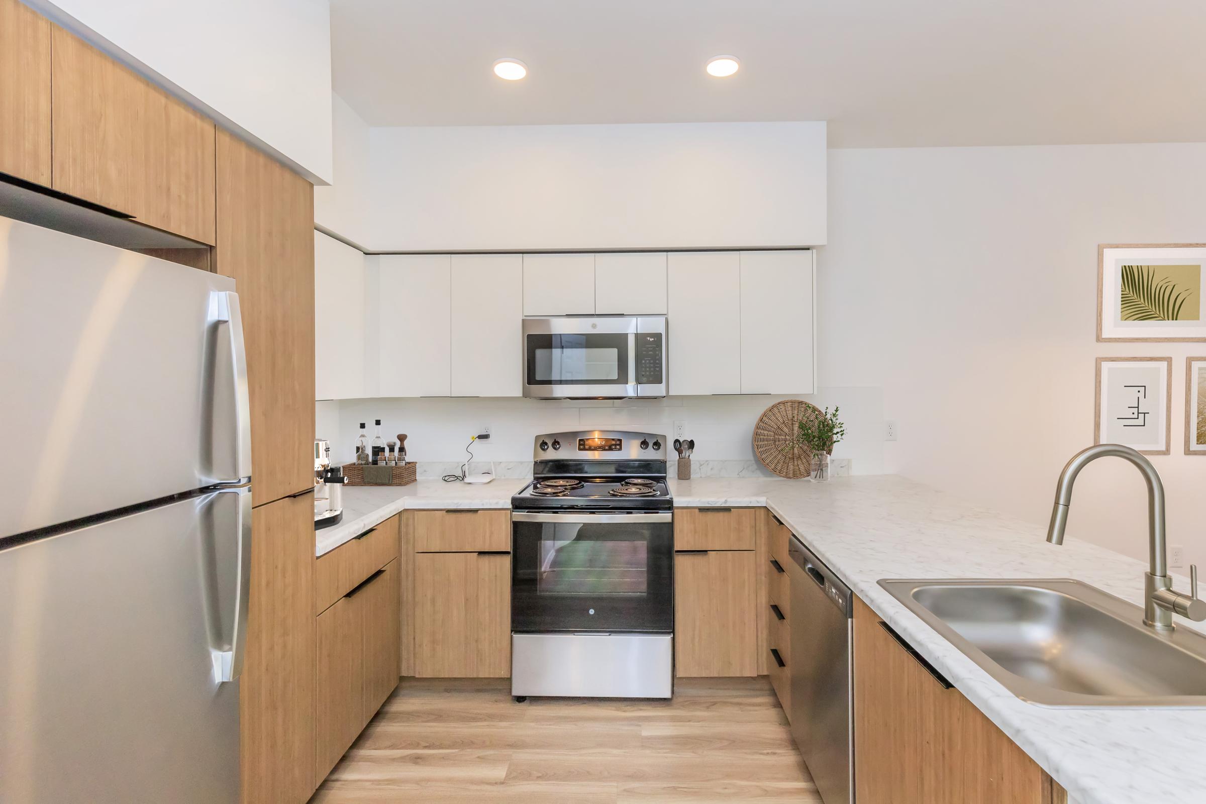 Modern kitchen featuring wood cabinetry and stainless steel appliances. It includes a refrigerator, oven, microwave, and a sink with a countertop. The space is well-lit with recessed lighting and has decorative accents on the walls, creating a clean and contemporary look.