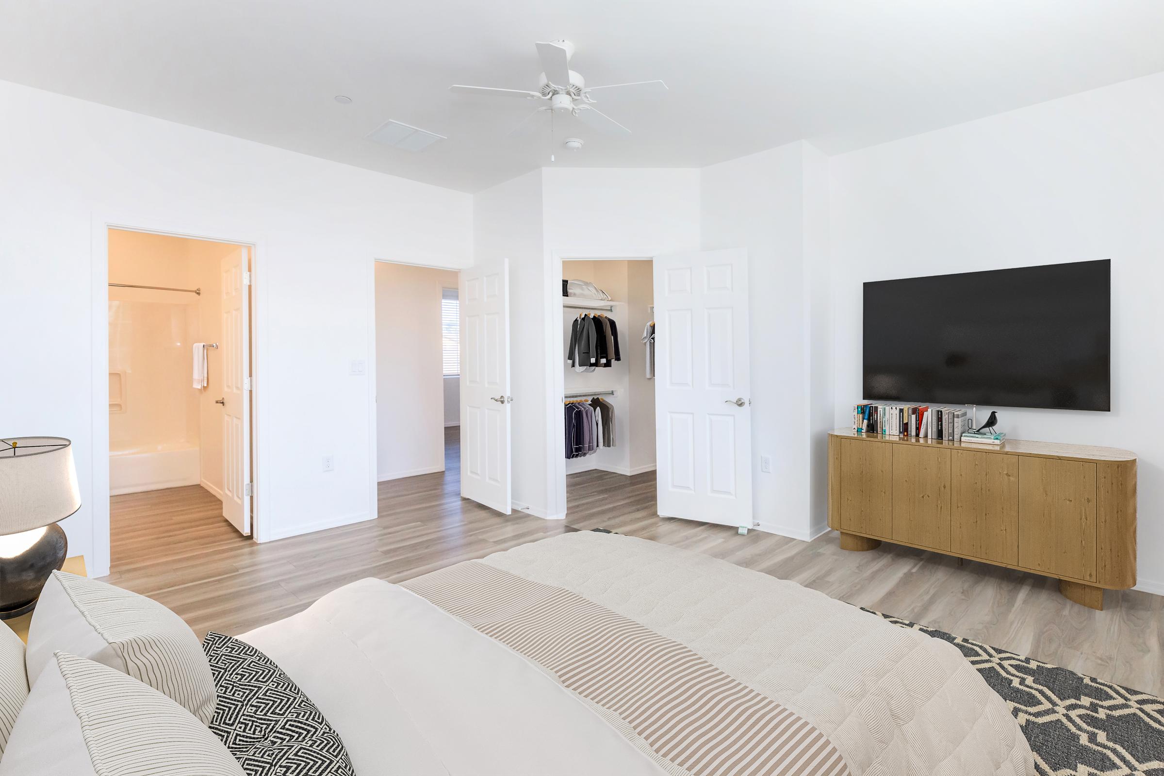 A modern bedroom featuring a large bed with neutral-colored bedding, a stylish area rug, and a bedside lamp. Opposite the bed, there’s a wall-mounted flat-screen TV above a wooden TV stand with books and decorative items. Two doors lead to an ensuite bathroom and a closet with hanging clothes.