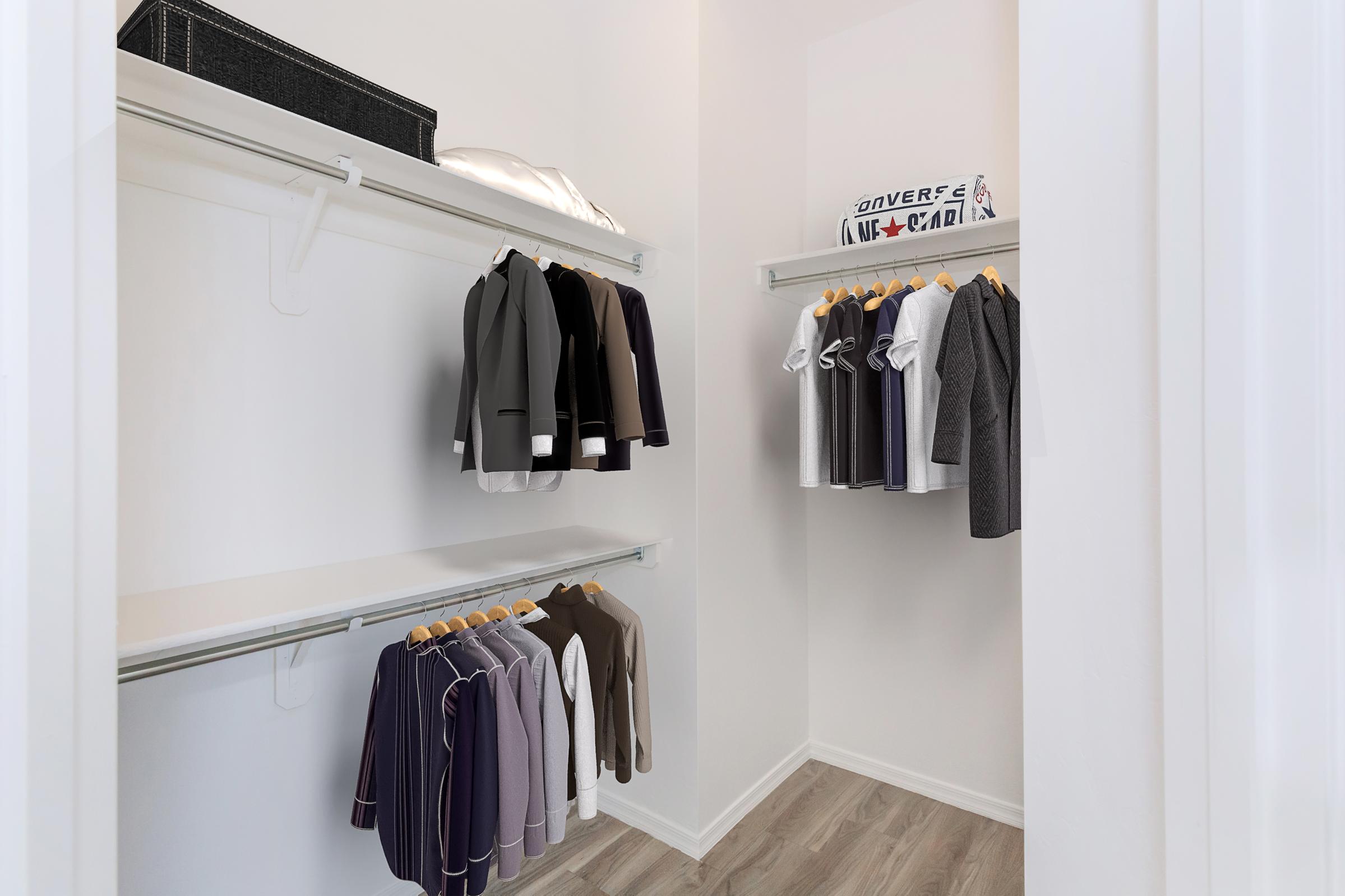 A well-organized closet featuring hanging clothes including formal suits and casual shirts. Shelves hold a black storage box and a stack of folded items, with light-colored walls and light wood flooring, creating a neat and spacious appearance.