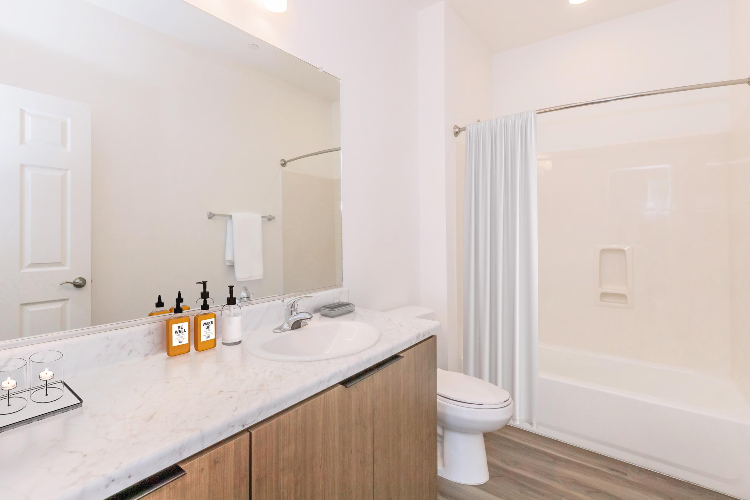 A bright and modern bathroom featuring a white bathtub with a shower curtain, a sleek vanity with a marble countertop, a sink, and several bottles of liquid soap. The space is well-lit, with a large mirror reflecting the room and a towel hanging on the wall.