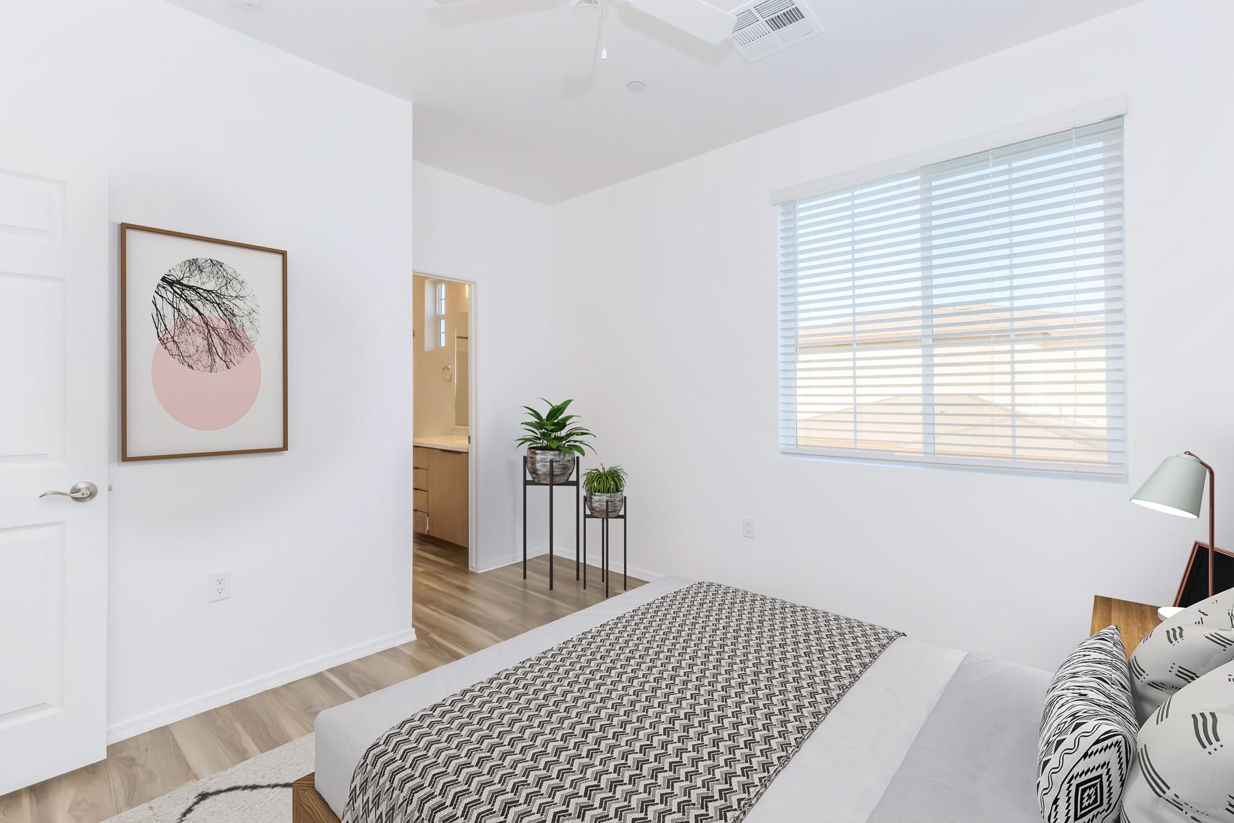 A modern, minimalist bedroom featuring a bed with a patterned gray and white duvet, a plant on a small side table, and a wall art piece with abstract design. The room has white walls, a large window with blinds allowing natural light, and a bathroom visible through an open door.