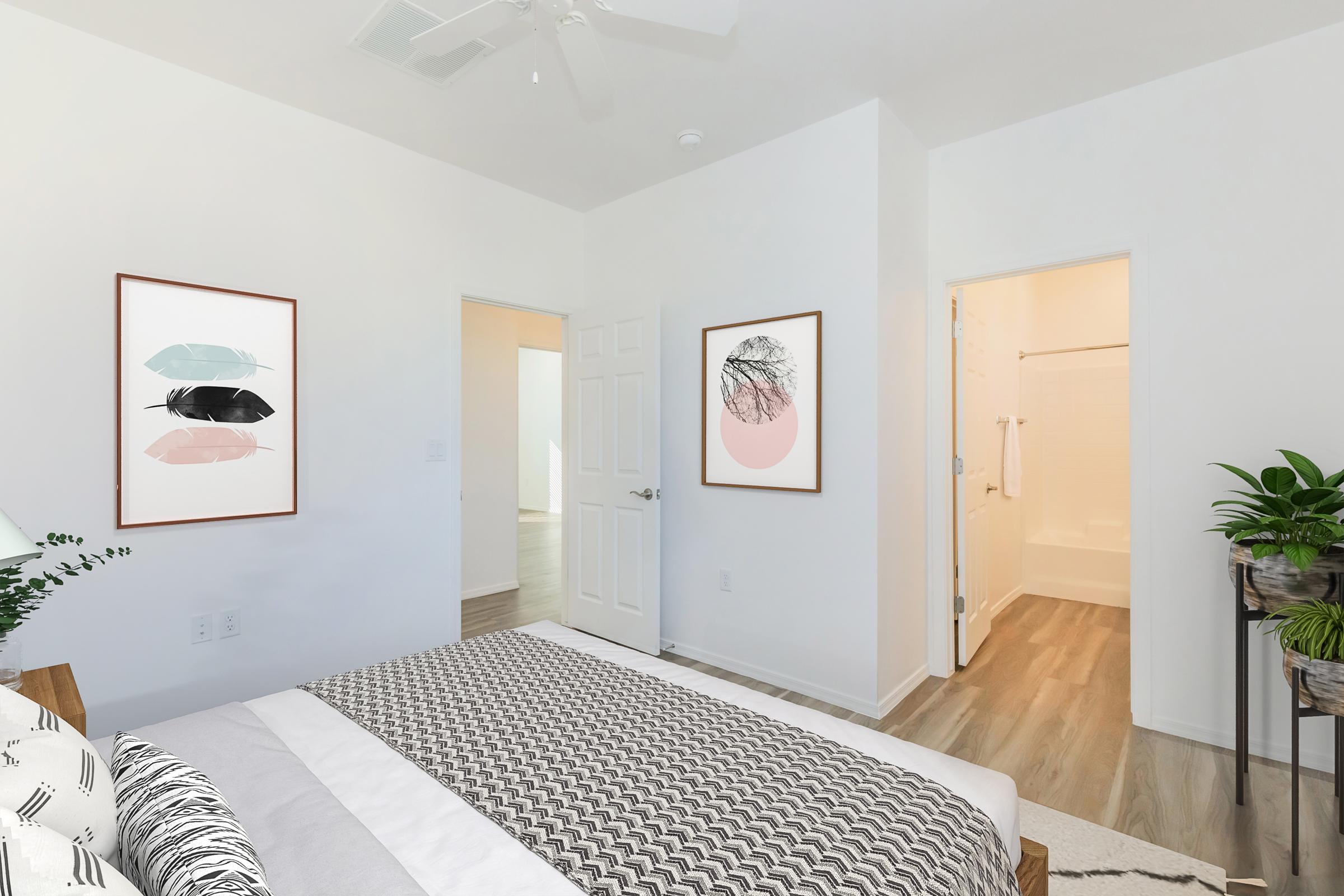 A modern bedroom featuring a bed with a patterned gray blanket, two framed art pieces on the white walls, and a potted plant. An open door leads to a bathroom, and another door opens to a bright space. The flooring is light wood, and the room has a minimalist design with neutral colors.
