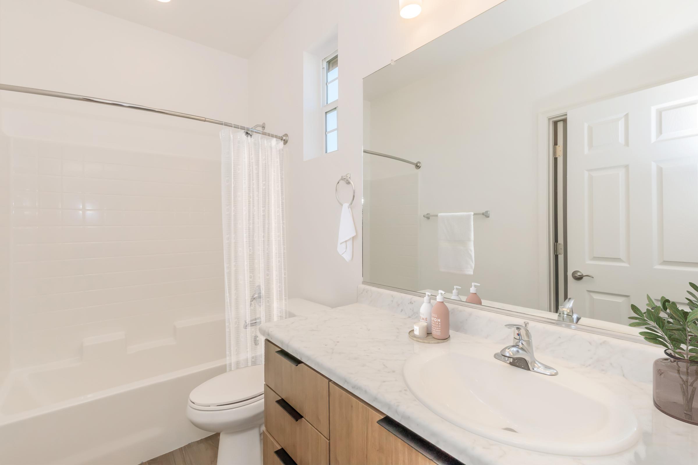 A modern bathroom featuring a white bathtub with a shower curtain, a marble countertop sink with soap dispensers, and a mirror. The walls are painted white, and there is a plant on the counter. A towel rack and a door leading to another room are visible. Bright lighting enhances the clean and airy feel.