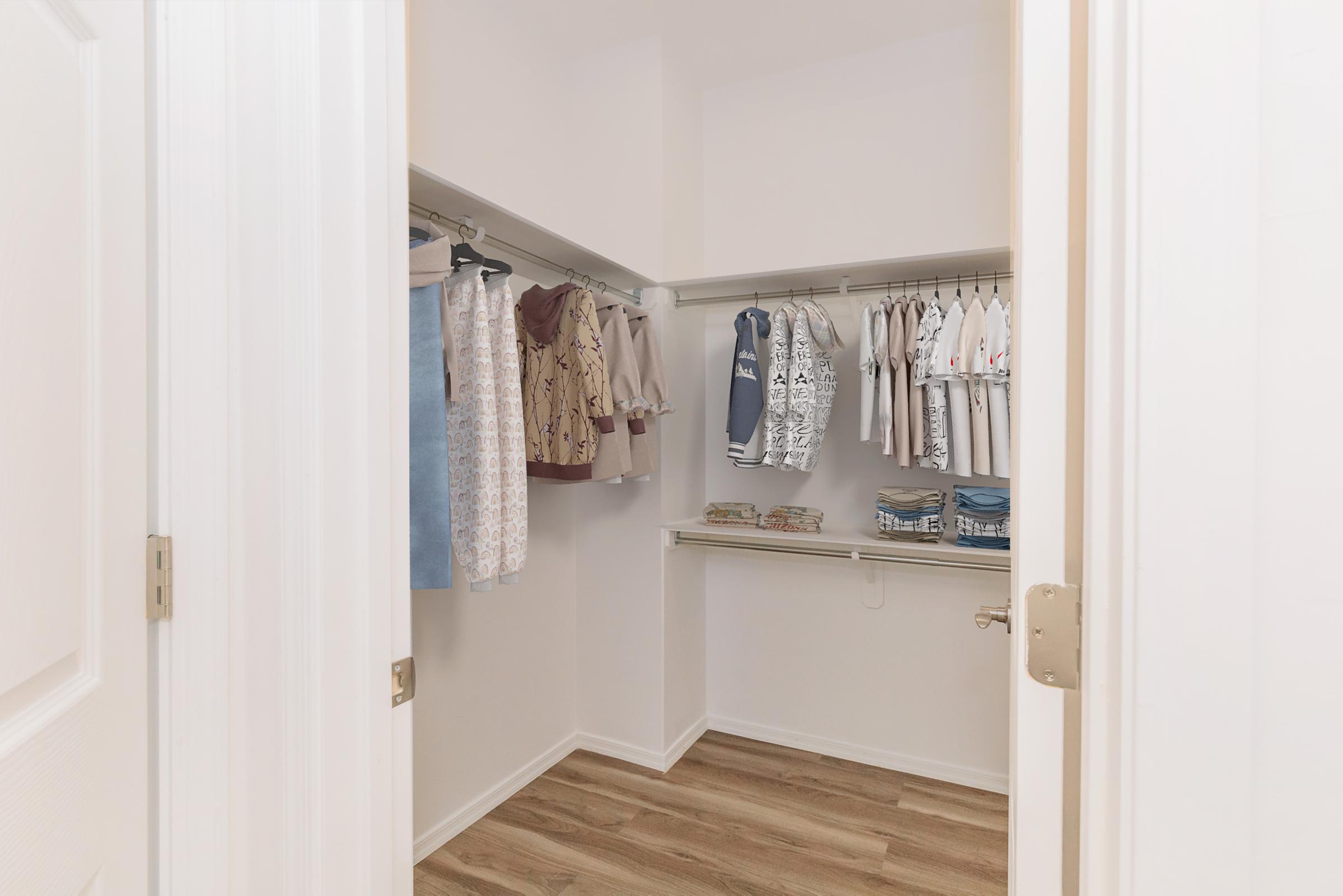 A clean, organized closet view from an open doorway. The closet contains neatly hung clothing items, including shirts and jackets, along with neatly folded stacks of clothes on a shelf. The floor features light wooden planks, and the walls are painted white, creating a bright and spacious appearance.