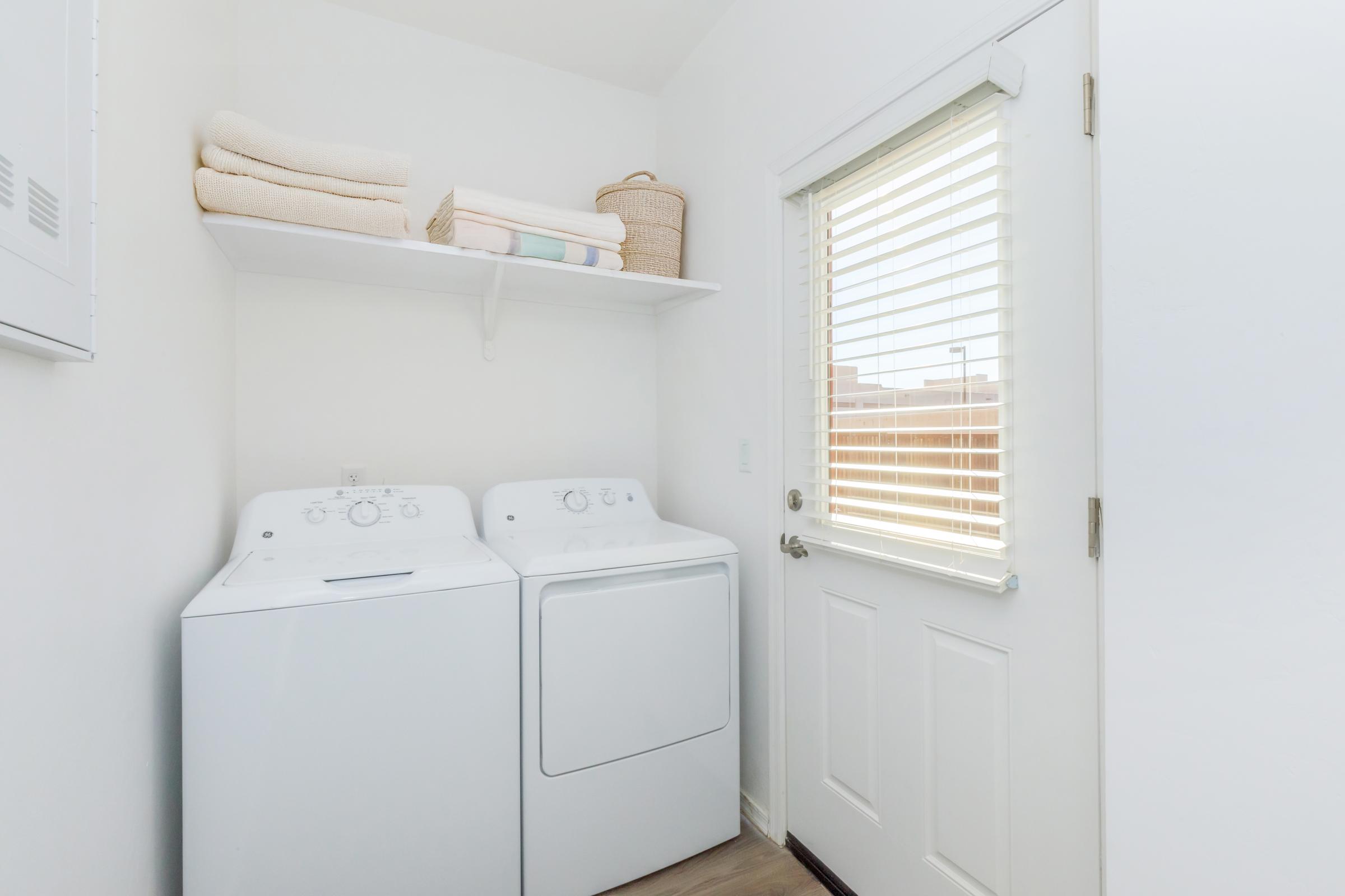 A clean laundry room featuring a white washing machine and dryer side by side. Above them, neatly folded towels are stacked on a shelf, and a woven basket is positioned next to them. A door with horizontal blinds allows natural light into the space. The walls are painted white, giving a bright, fresh appearance.
