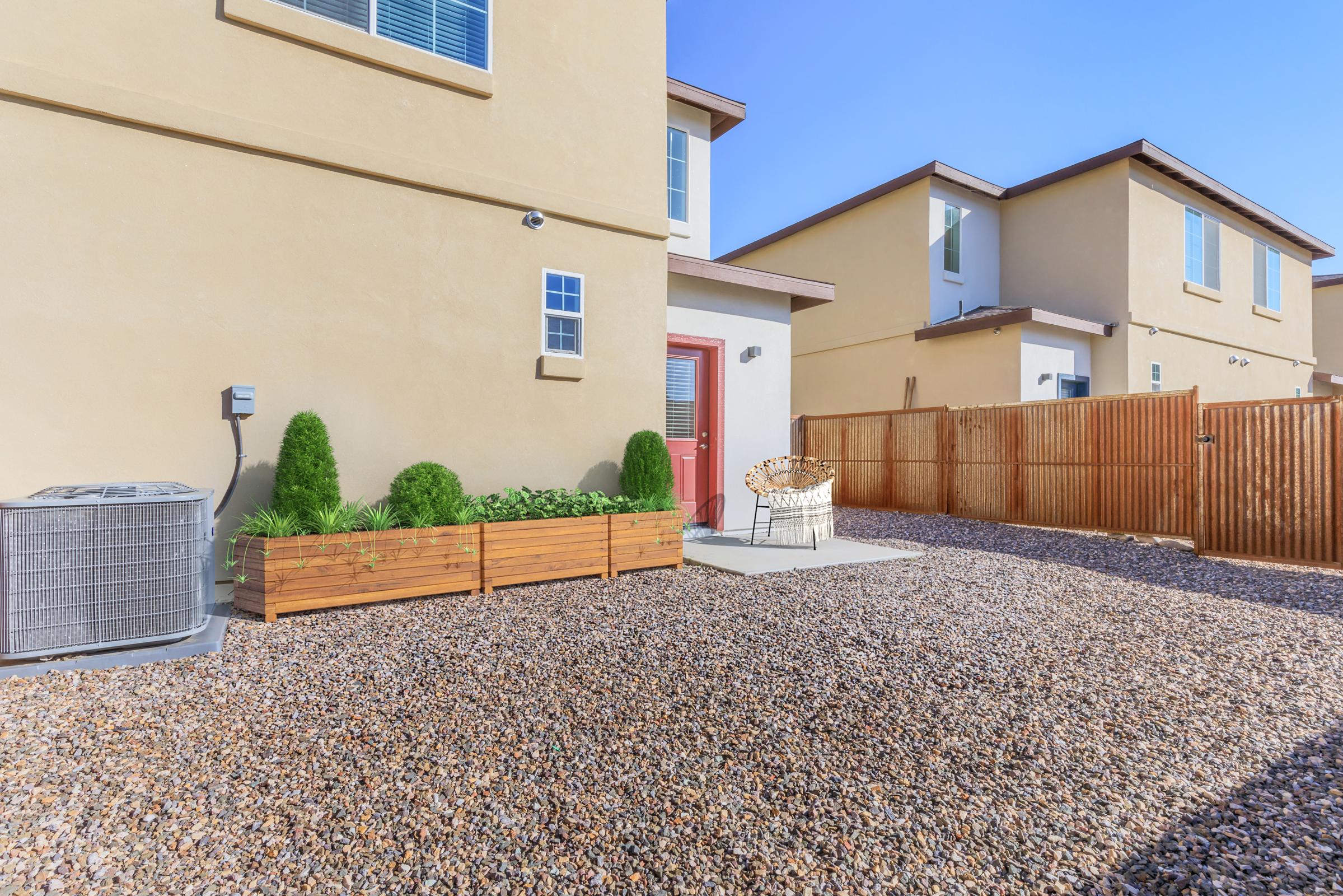 A small outdoor space featuring two houses, a wooden fence, a gravel surface, and a wooden planter with green shrubs. There is an air conditioning unit on the left and a decorative chair on a stone slab in the center, with clear blue skies above.
